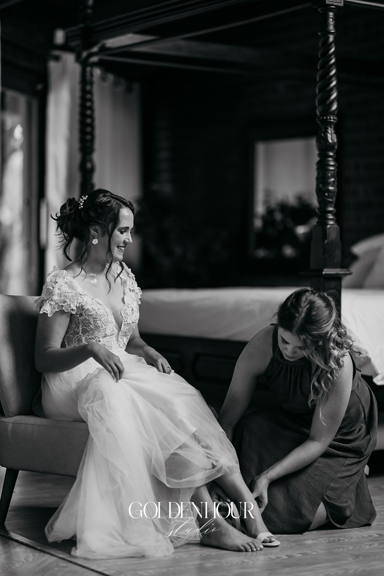 Black and white photo of smiling bride in lace gown having shoes put on by bridesmaid indoors.