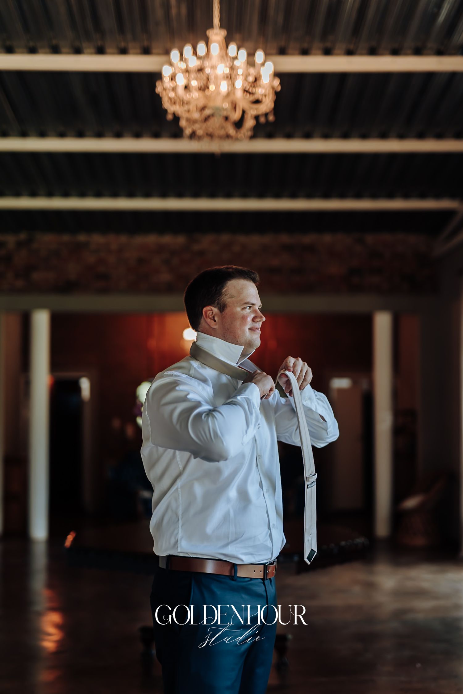Groom adjusting his white tie under a chandelier in a dimly lit wedding venue.
