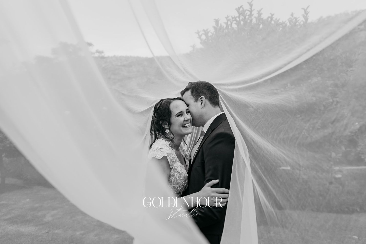 Black and white romantic photo of newlyweds embracing beneath billowing wedding veil.