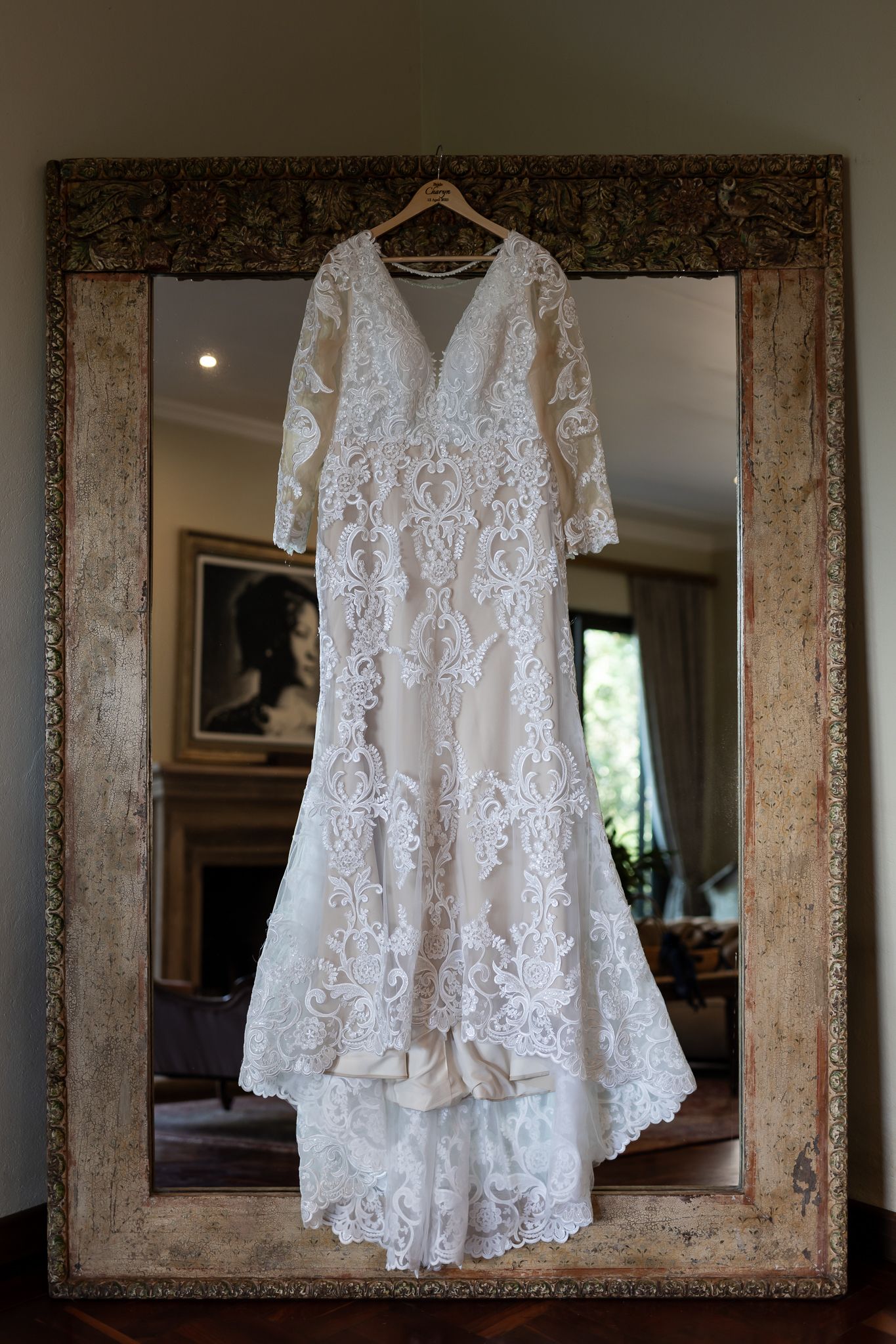 Elegant lace wedding dress with long sleeves displayed on hanger against ornate vintage mirror.