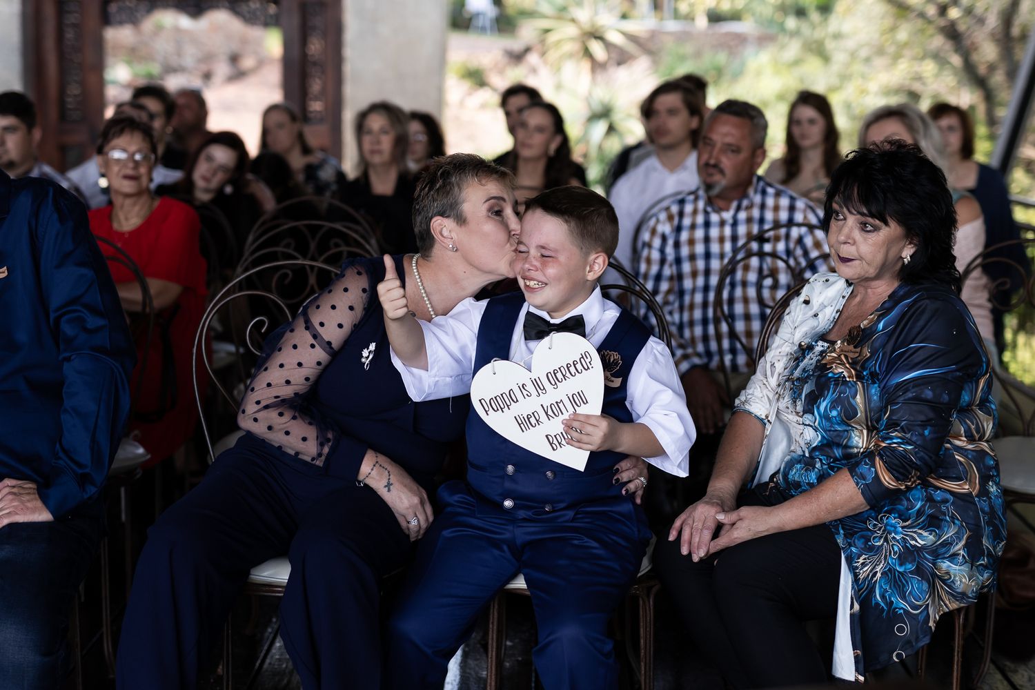 Wedding guests gather around a young ring bearer holding a heart-shaped sign.