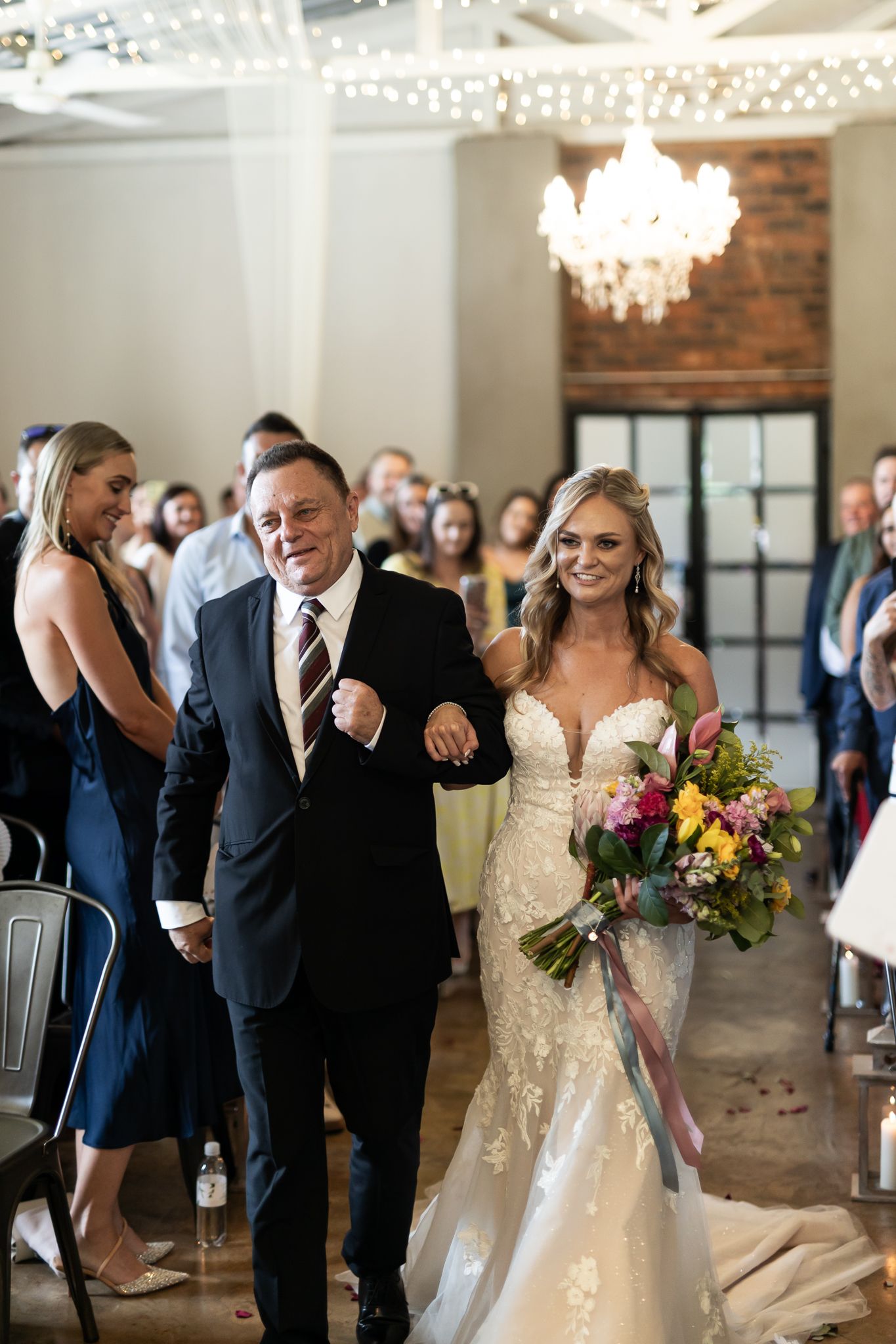 A bride in a lace gown walks down the aisle with her escort under elegant chandeliers.