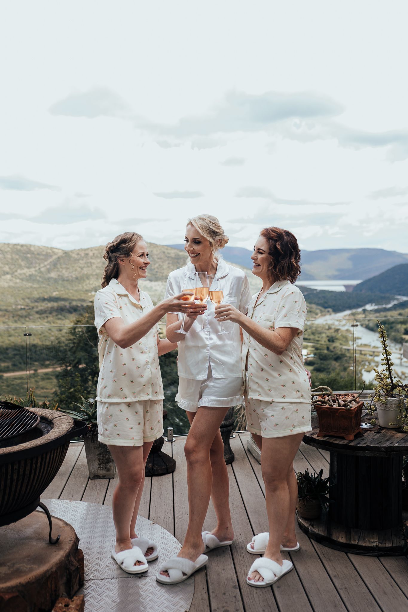 Three people in white loungewear celebrating with drinks on mountain view terrace.
