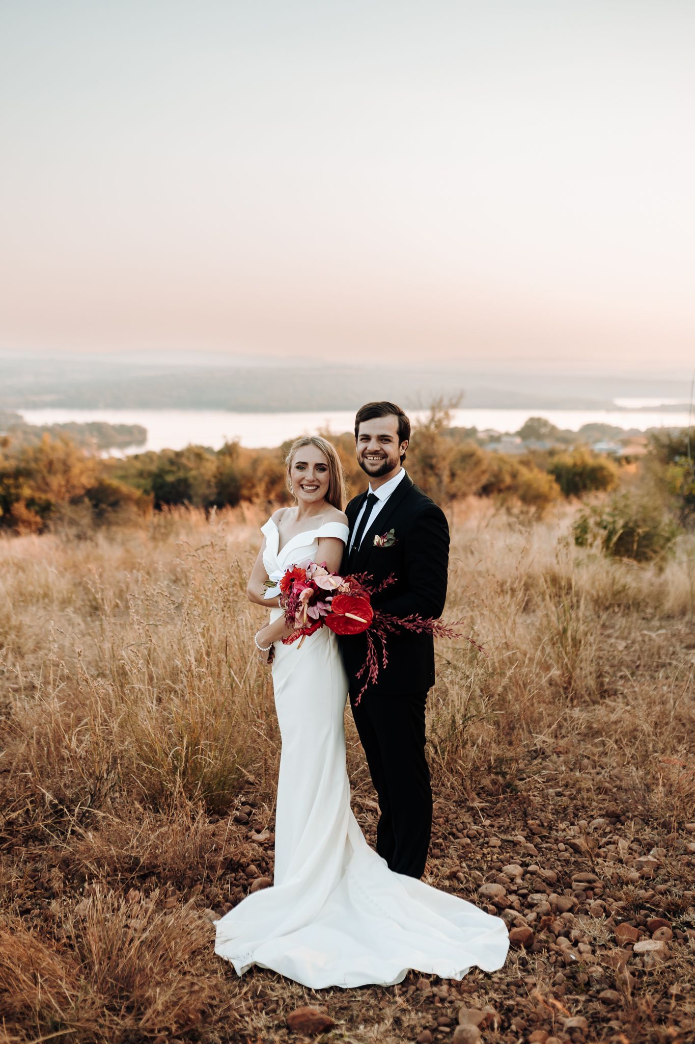 A newlywed couple poses on a hilltop overlooking a lake at dusk.