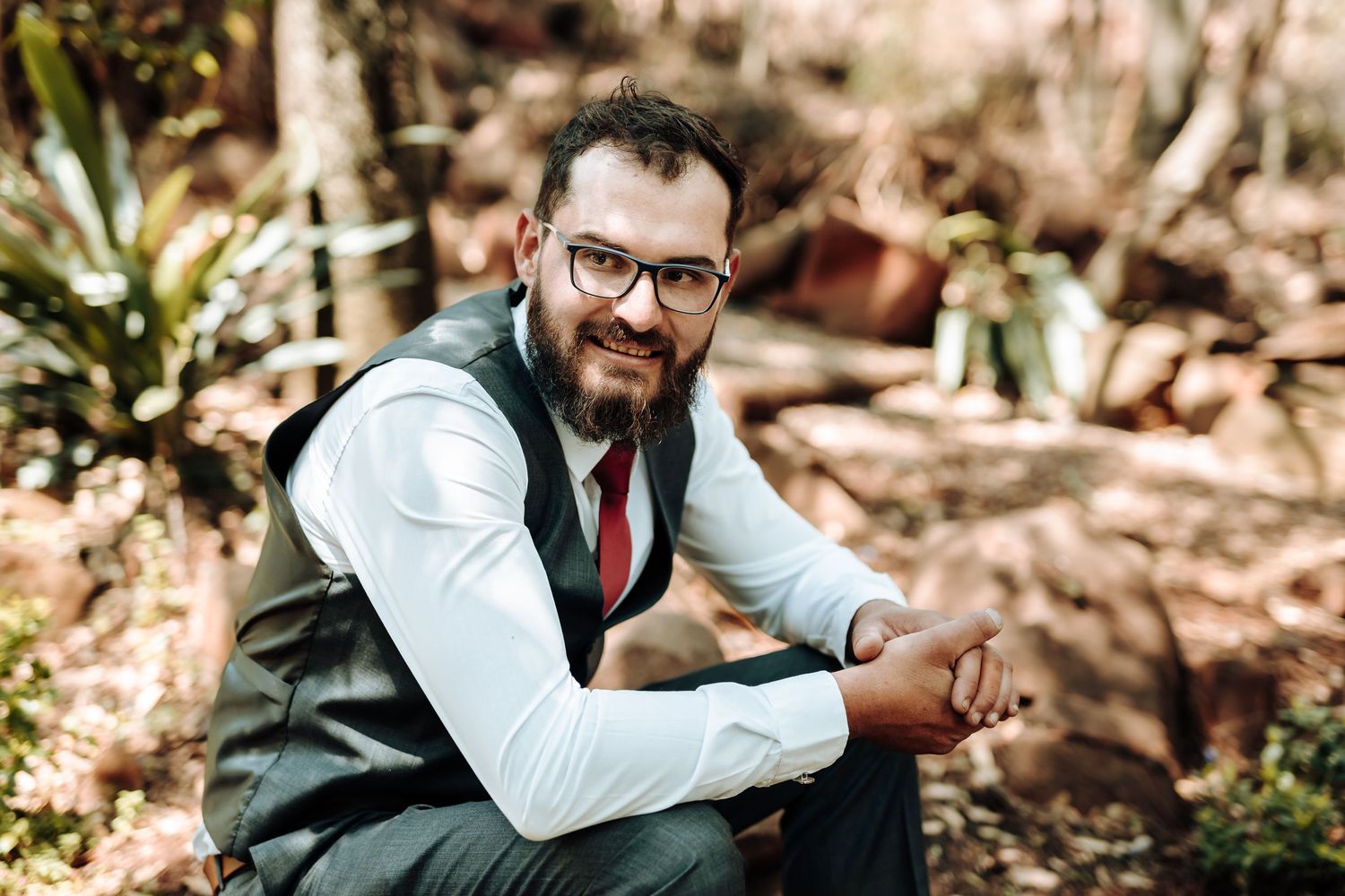 Portrait of groomsman in formal vest and red tie seated in outdoor garden setting.