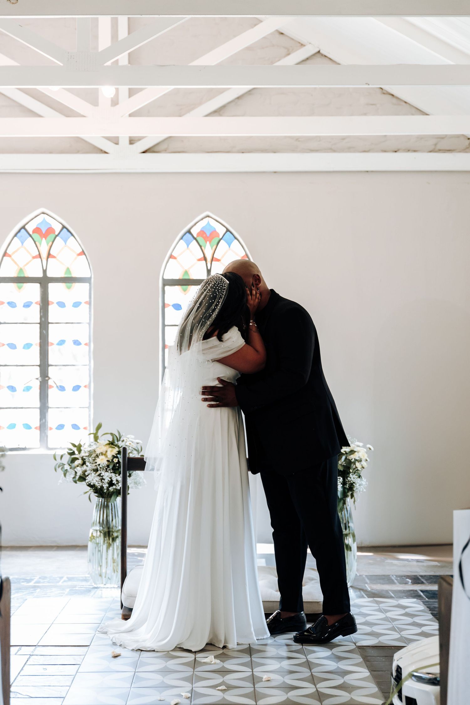 A tender moment between newlyweds in a chapel with stained glass windows and white beams overhead.