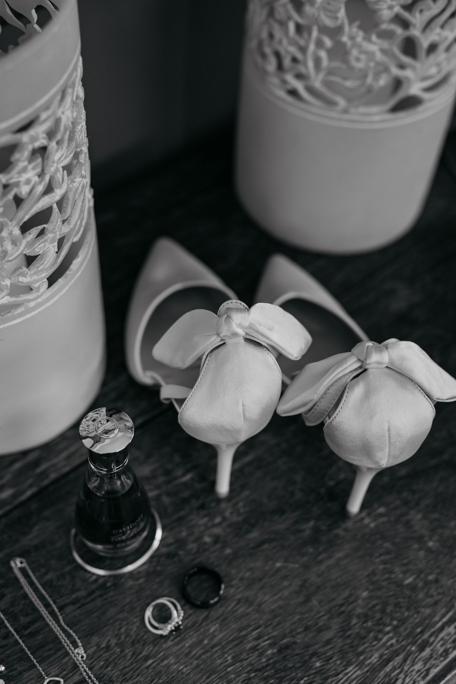 White bridal heels with bow details next to decorative vases and wedding rings on dark surface.