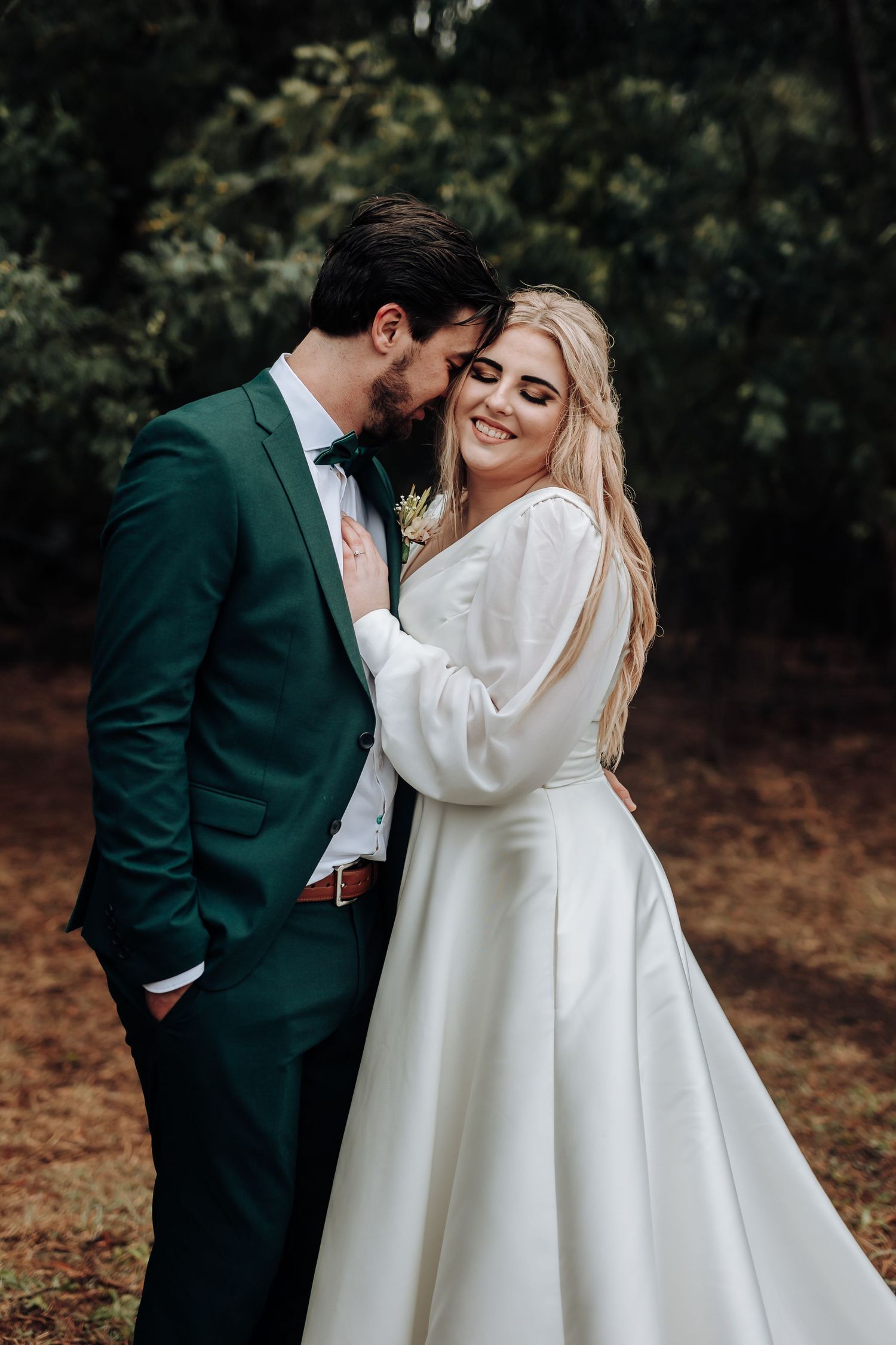 Romantic forest wedding portrait with couple in emerald suit and flowing white dress sharing intimate moment.