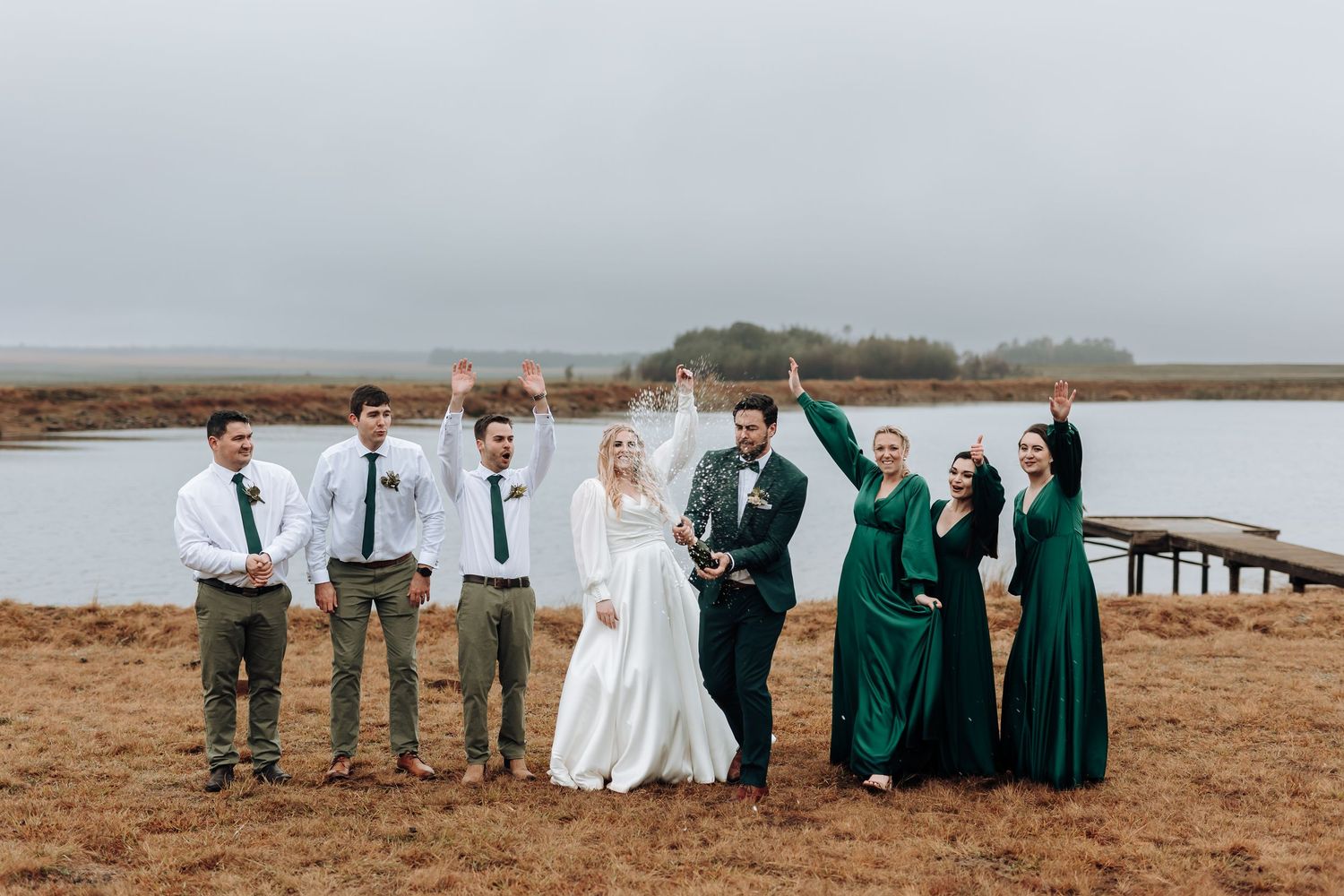 Wedding group formal portrait with emerald dresses and white shirts against water backdrop.