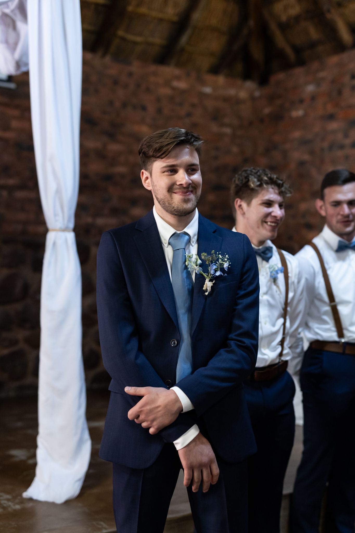 Groomsmen in formal attire stand together inside rustic brick venue during wedding ceremony.