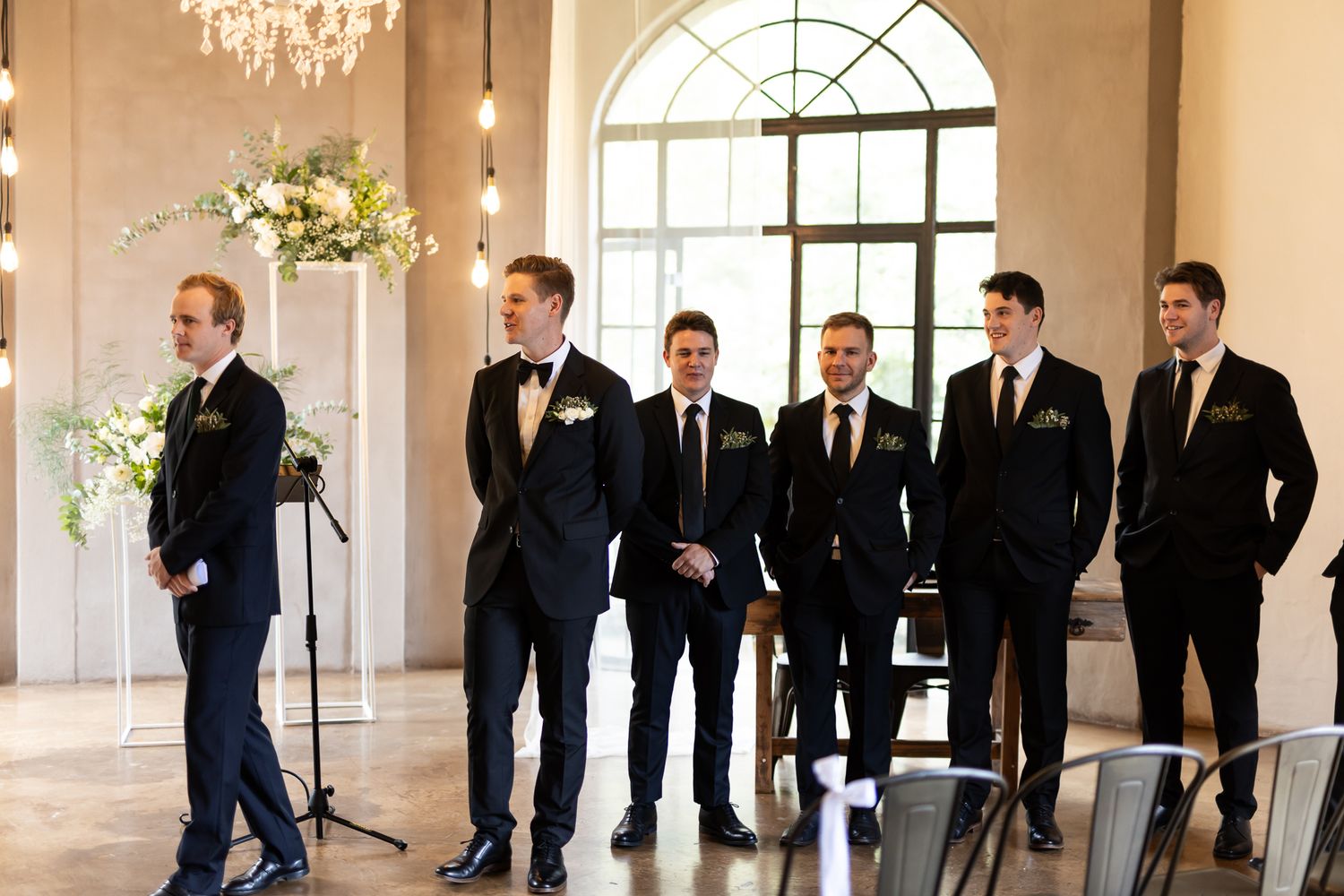 Groomsmen in dark suits stand together in an elegant venue with arched windows and floral arrangements.
