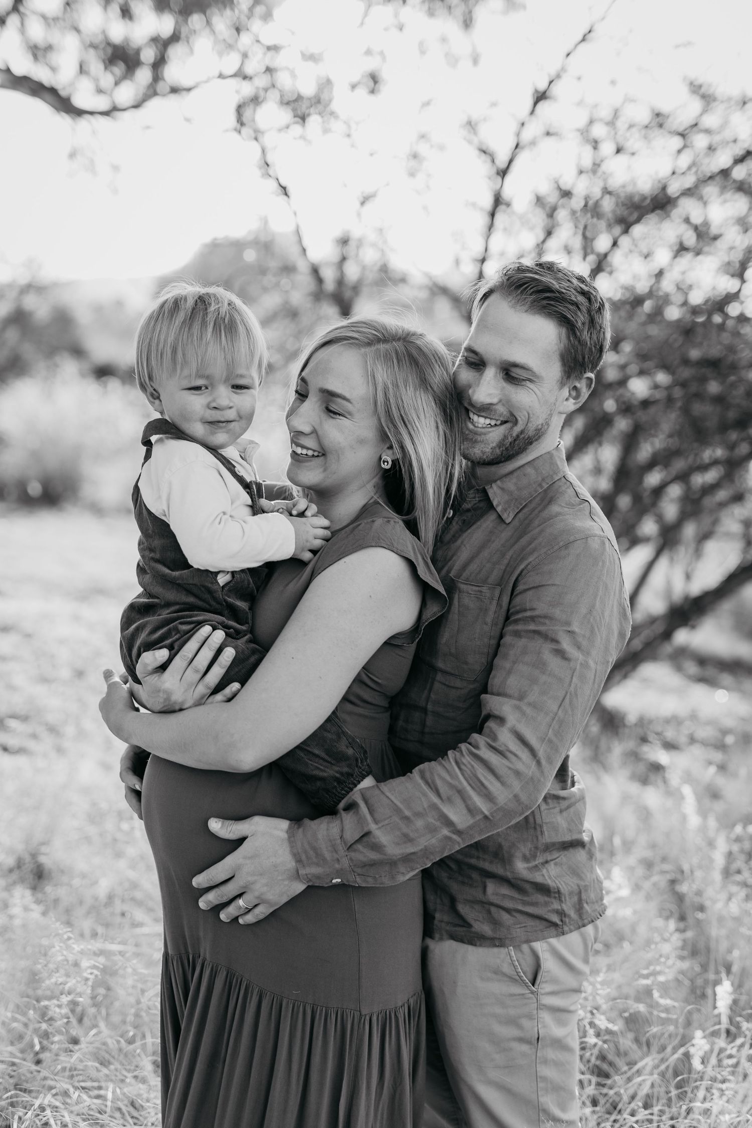 A family shares an intimate moment outdoors in black and white, embracing and smiling together in a natural setting.