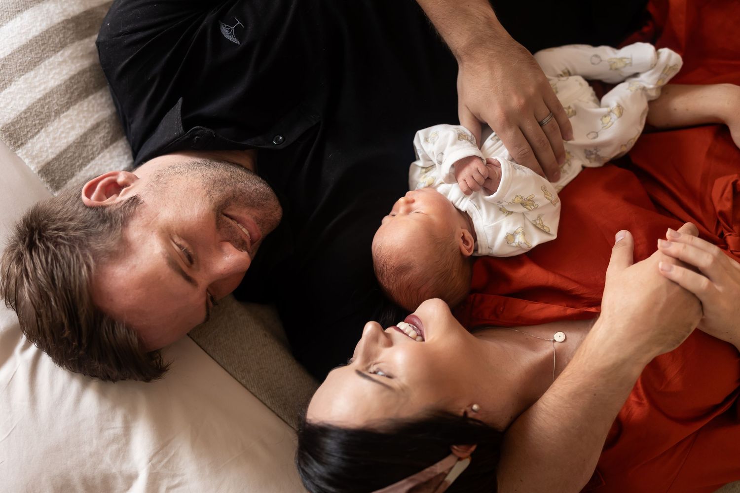 Hands tenderly comfort a sleeping newborn wrapped in a white blanket against a red blanket.