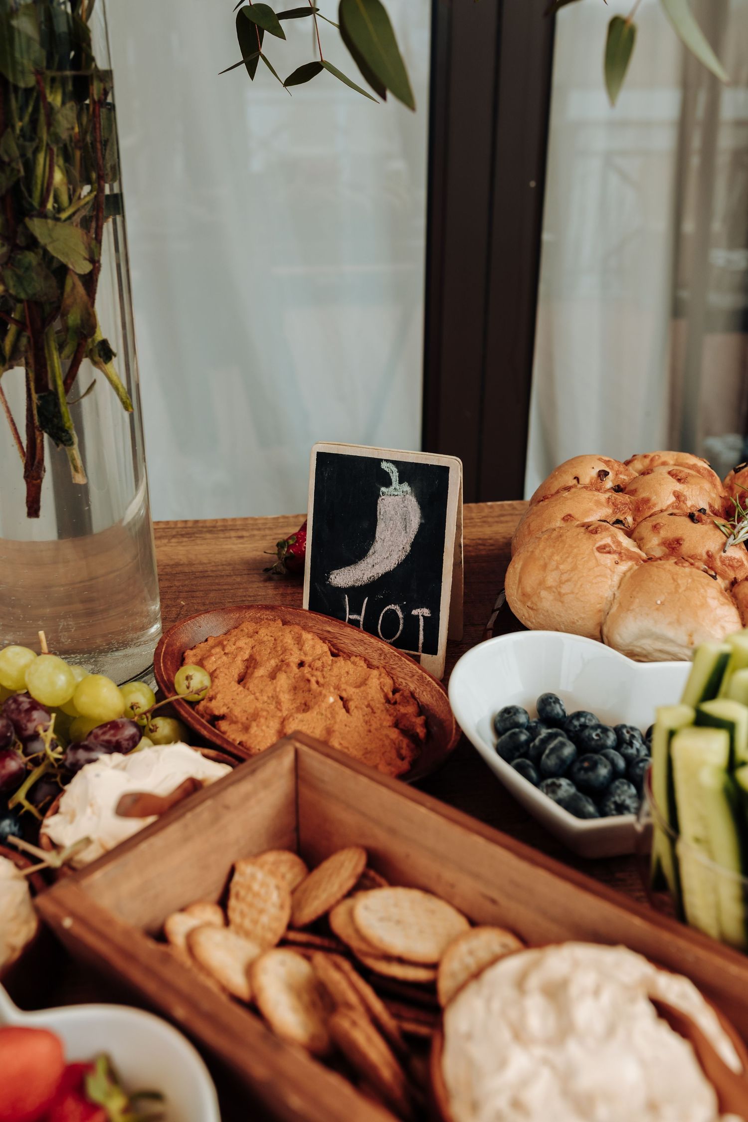 A rustic food spread with blueberries, crackers, bread, and fresh vegetables arranged on a wooden surface with a small chalkboard sign.