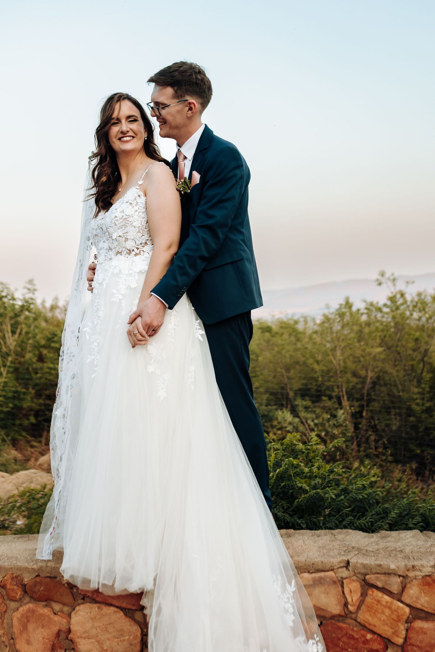 A romantic wedding couple embraces outdoors at sunset on a rocky overlook surrounded by greenery.