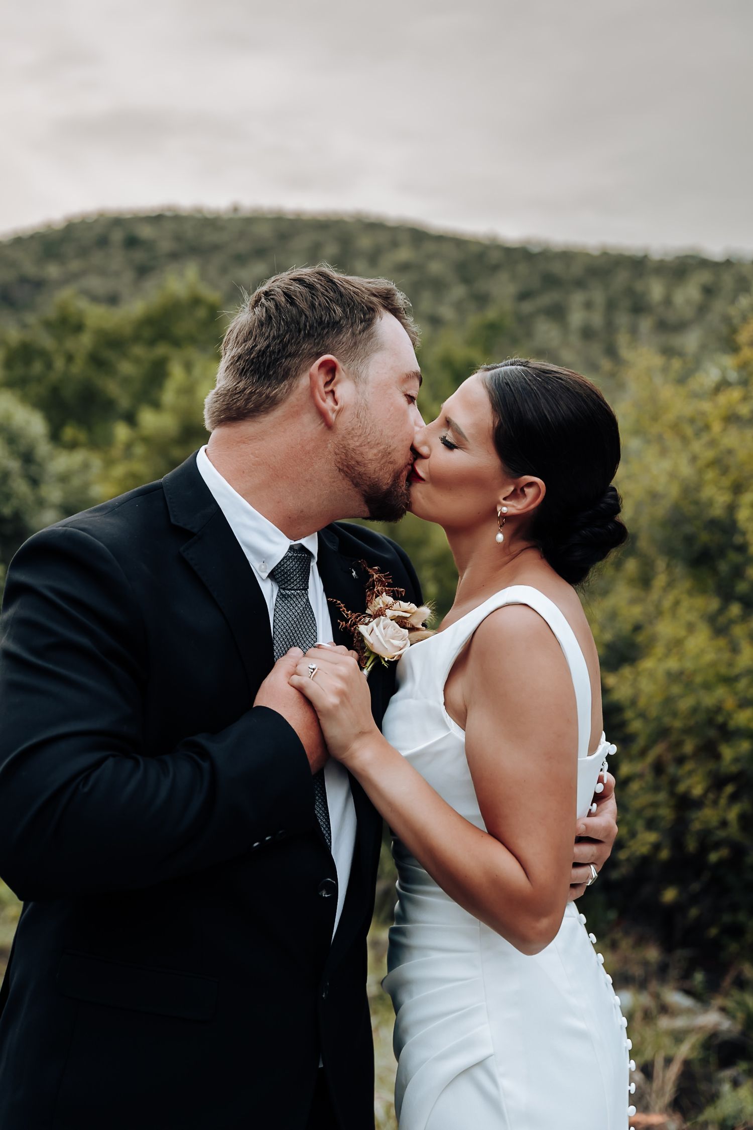 A couple shares a romantic kiss against a backdrop of lush green hills and trees.
