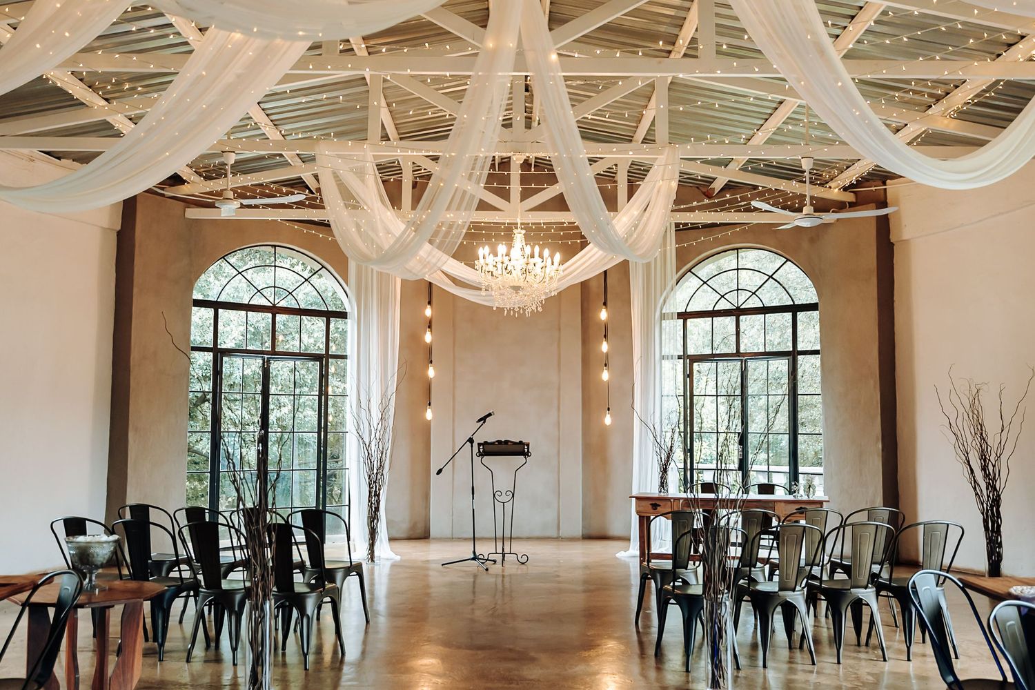 Elegant white wedding venue interior with draped fabric ceiling, large windows, and black chairs arranged for ceremony.