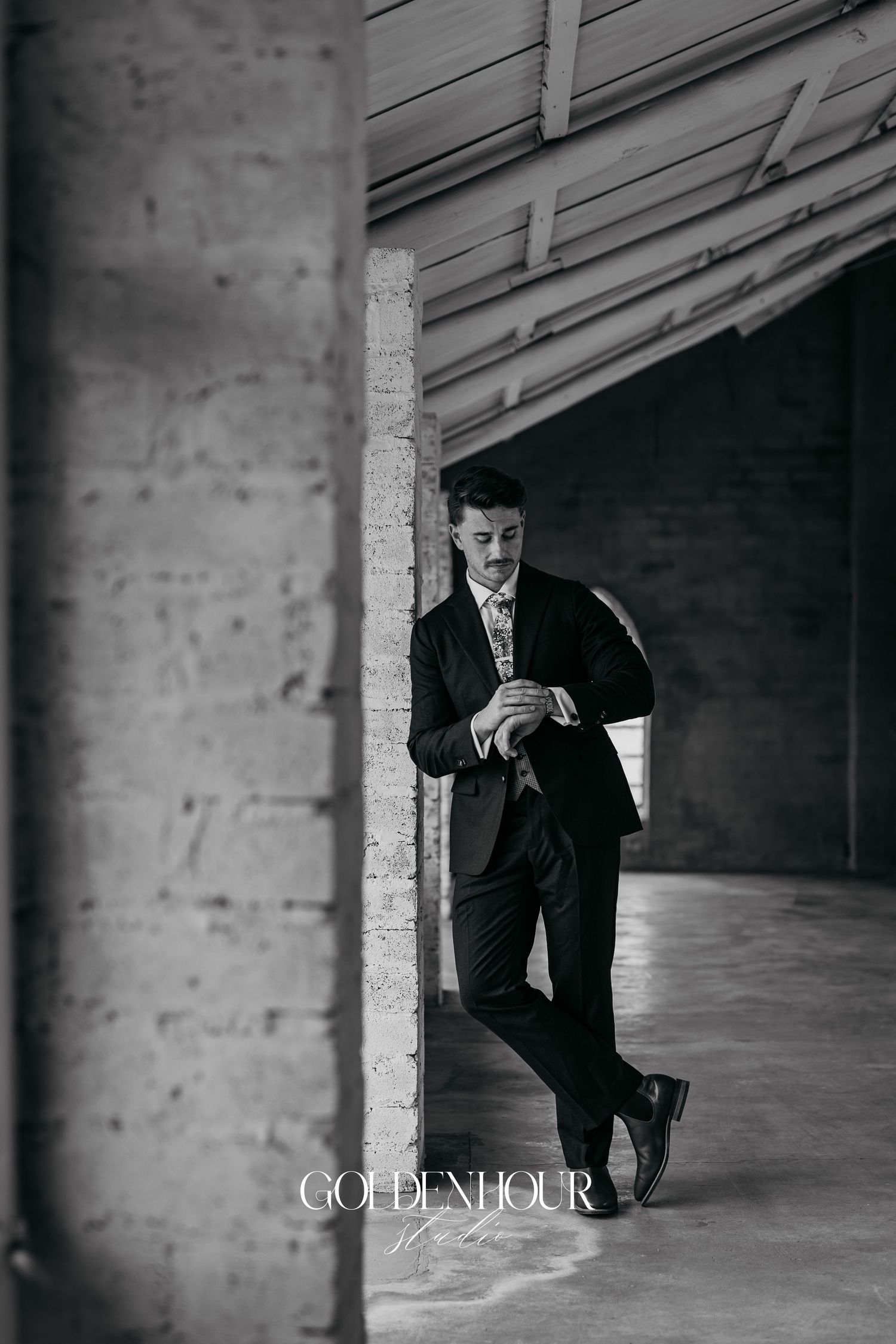 Moody black and white photos of a person in a suit standing against a brick wall in an industrial space.
