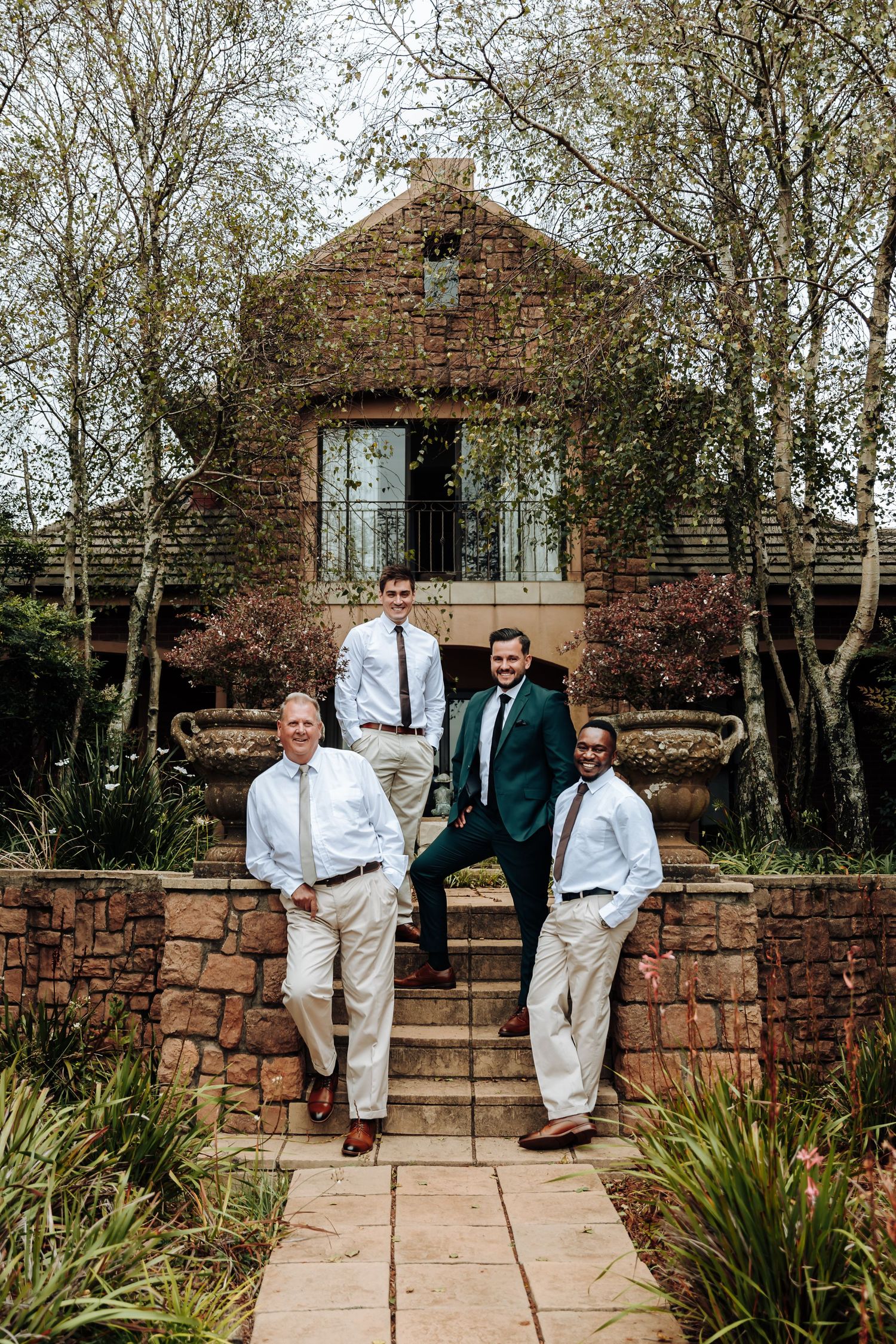 Three people sitting and standing on stone steps outside a rustic building surrounded by trees and greenery.