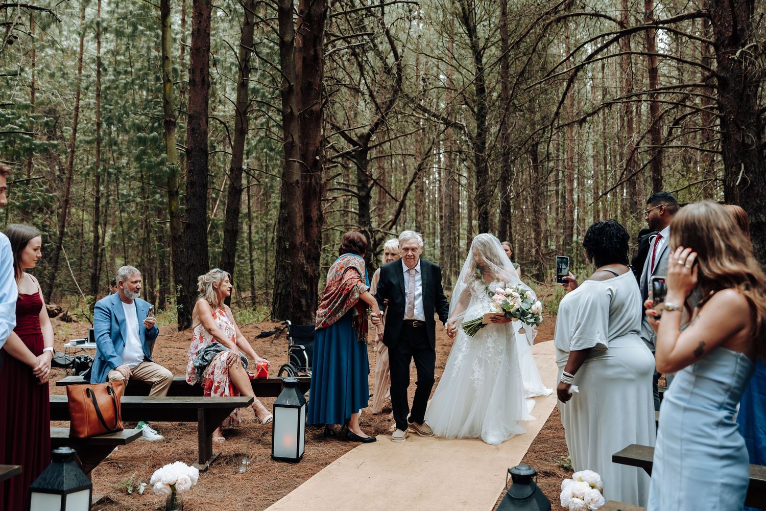 A small intimate outdoor wedding ceremony takes place on a wooden walkway surrounded by tall pine trees in the forest.