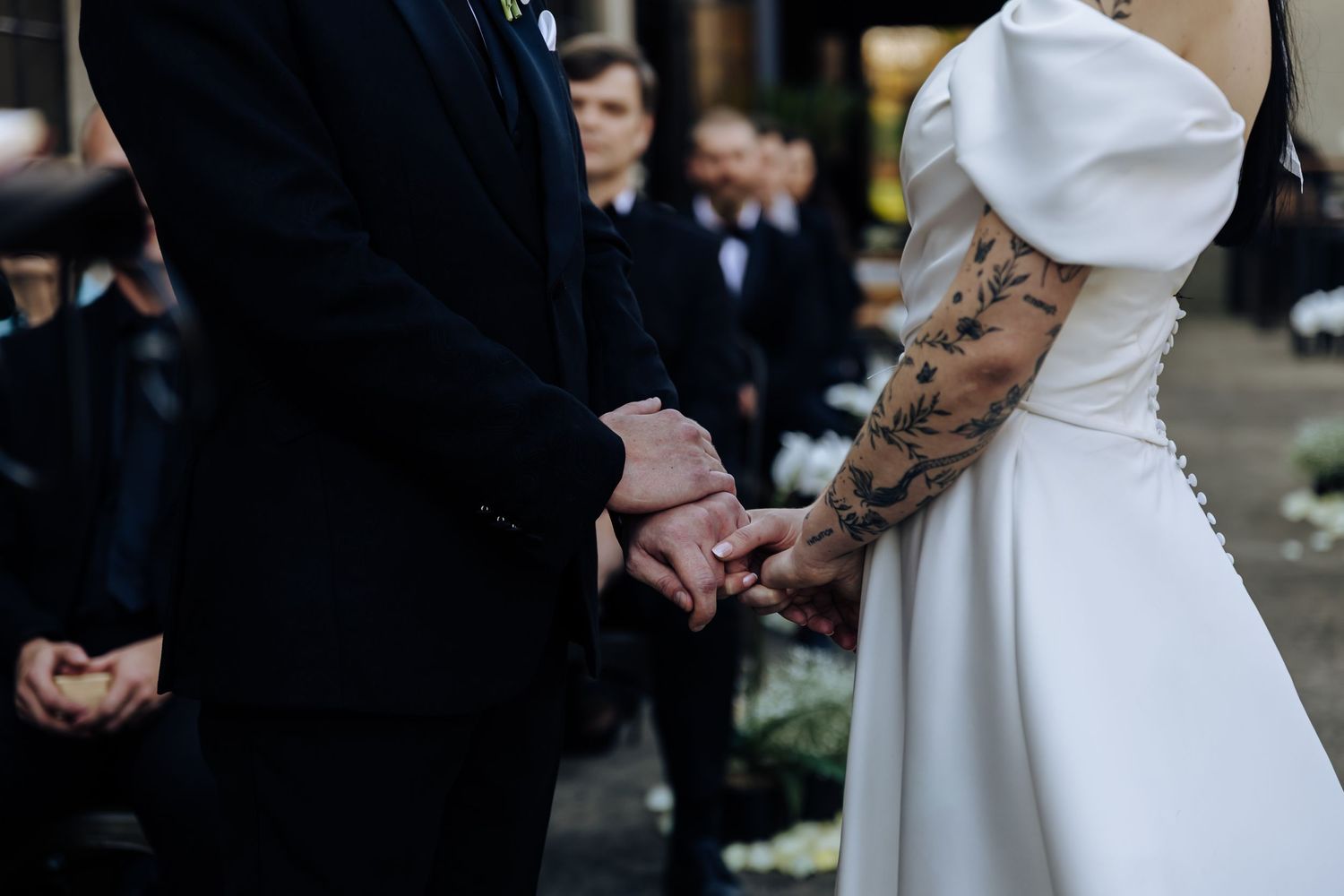 A close-up shot shows a bride in a white dress and groom in black holding hands during an outdoor wedding ceremony.
