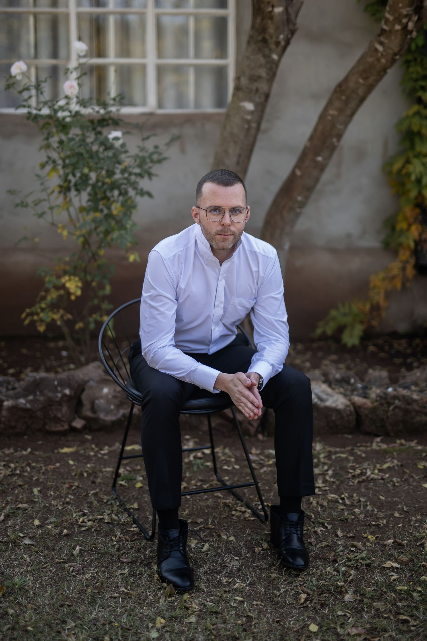 Professional seated in white shirt and dark pants in outdoor garden setting with stone wall and greenery in background.