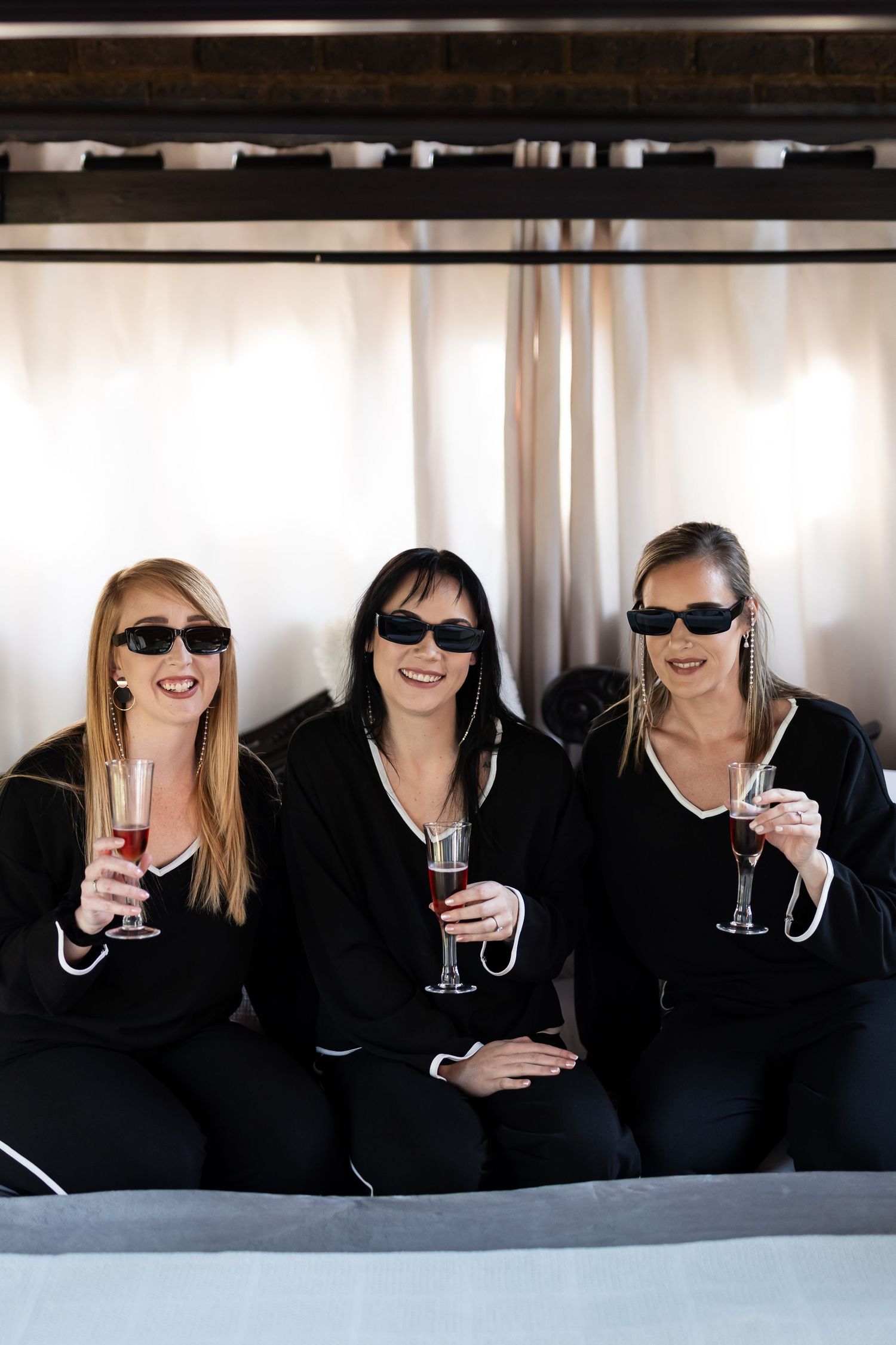 Three friends in matching black outfits and sunglasses smile while holding wine glasses together during a celebration.