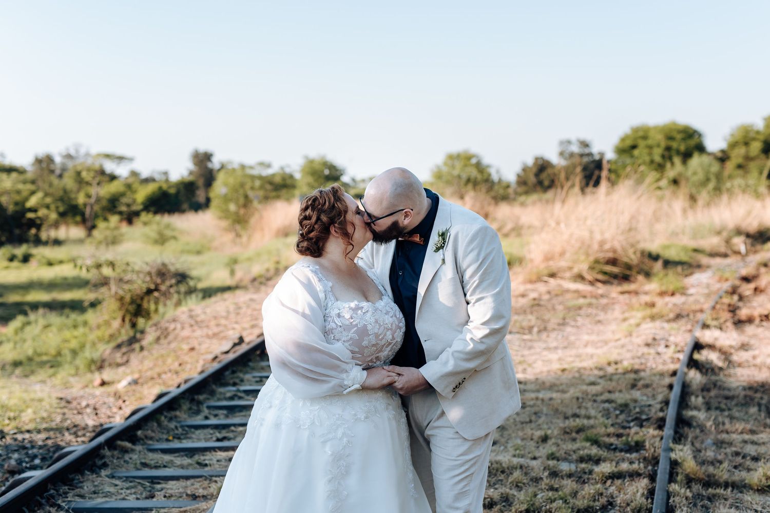 A romantic moment between newlyweds in white attire standing on rustic train tracks surrounded by nature.