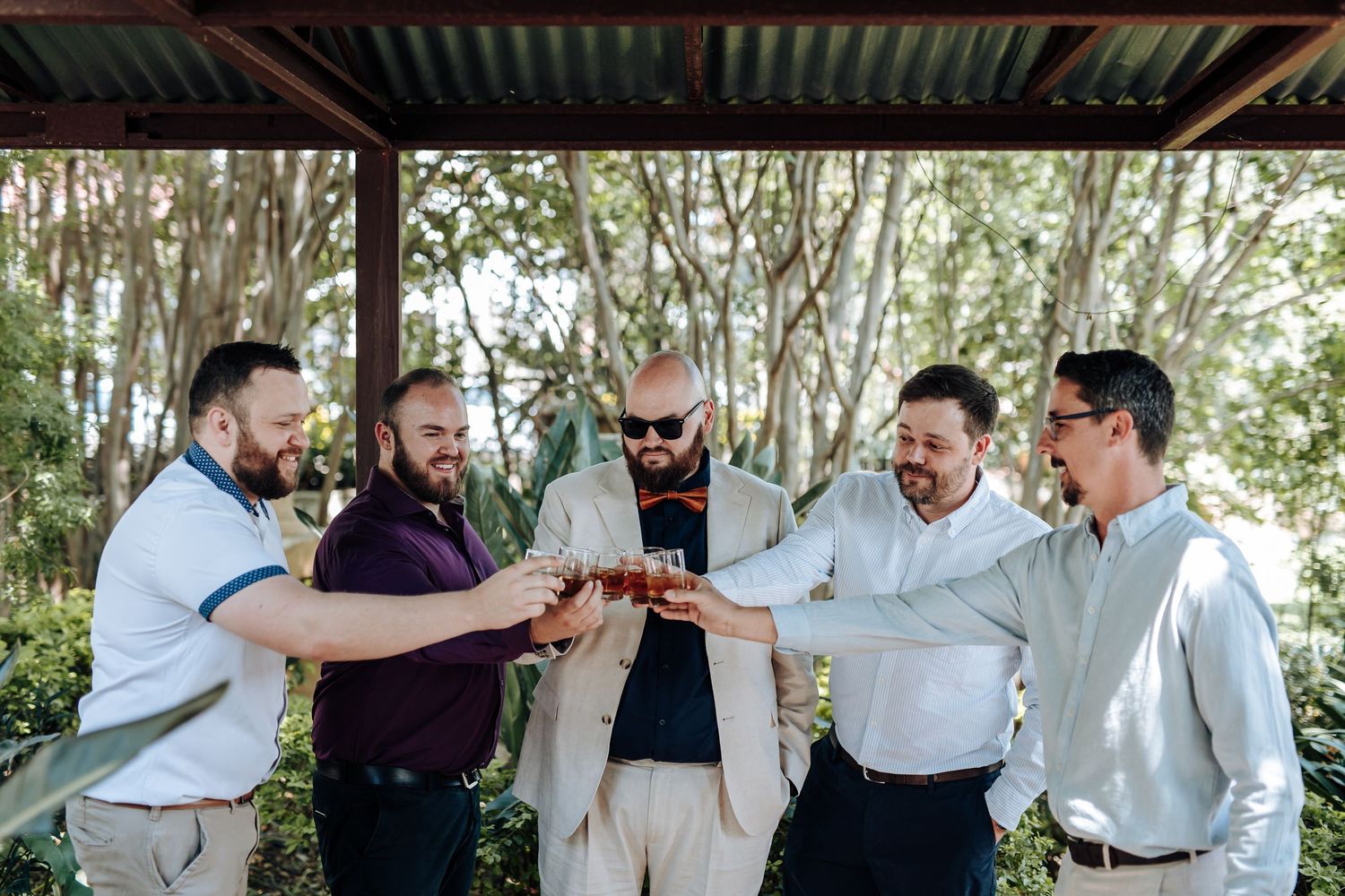 A group of groomsmen share a celebratory drink and laugh together in a rustic outdoor setting.