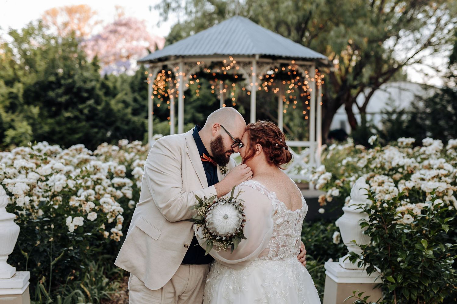 A couple shares an intimate moment in a blooming garden under a cloudy sky.