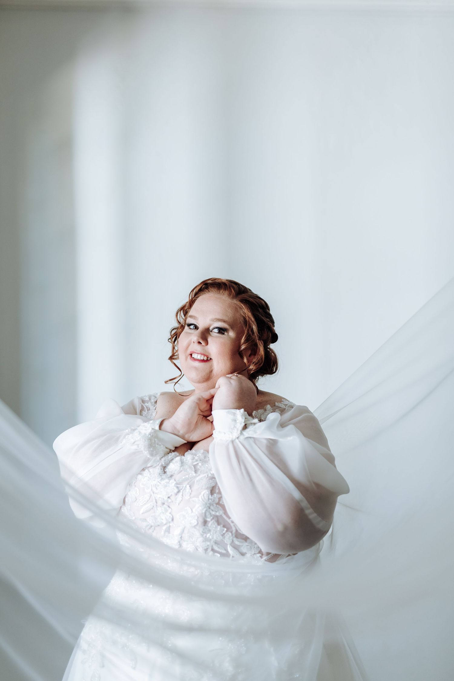 A figure wrapped in flowing white fabric poses gracefully in a bright minimalist setting.