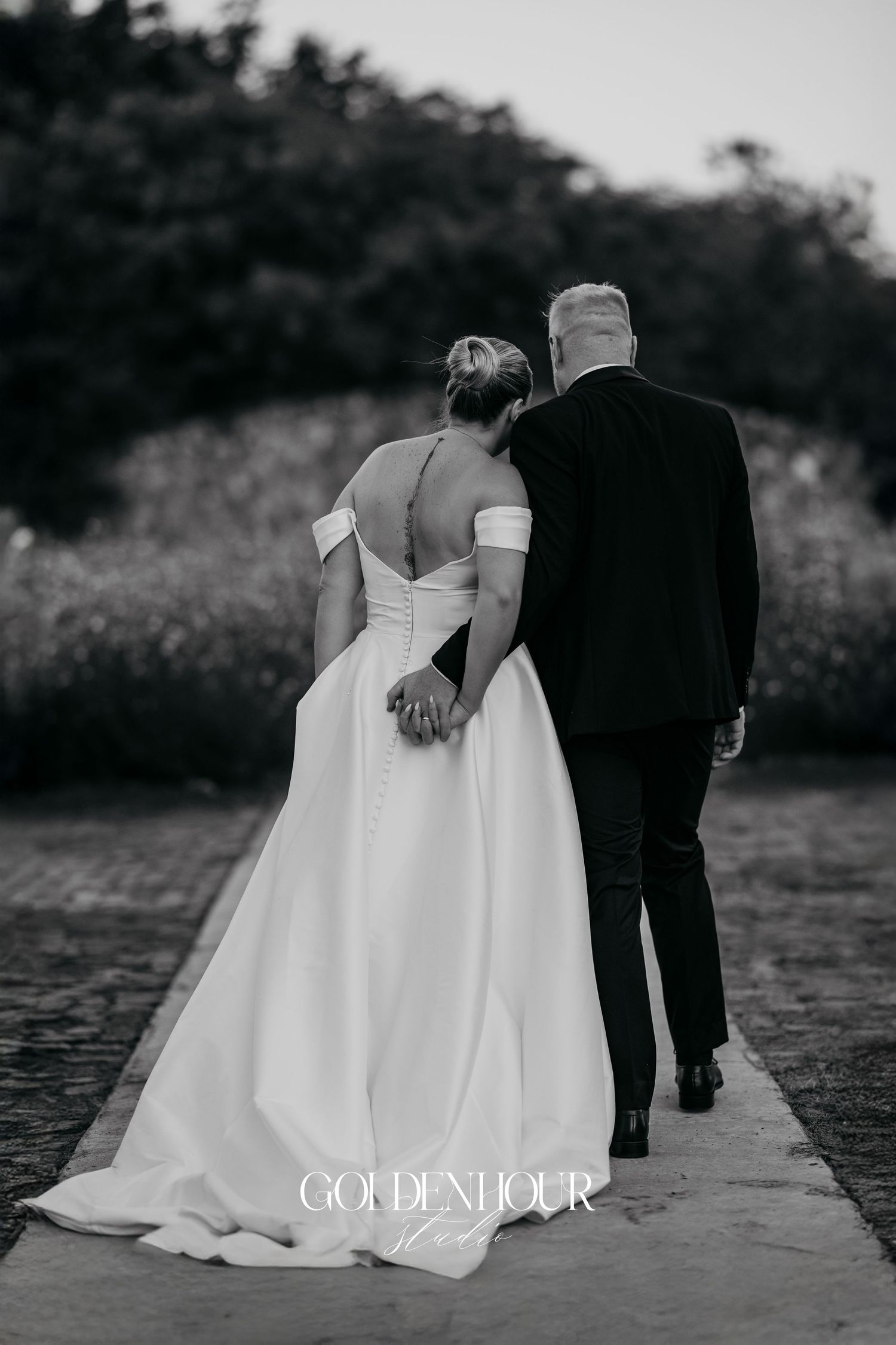 Black and white wedding photo showing couple from behind, bride in off-shoulder gown and groom in suit walking outdoors.