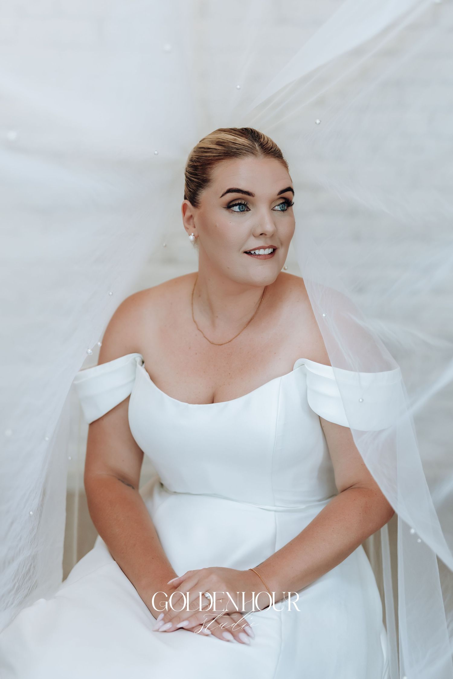 A bride in an elegant white off-shoulder wedding gown poses against a light background for bridal portraits.