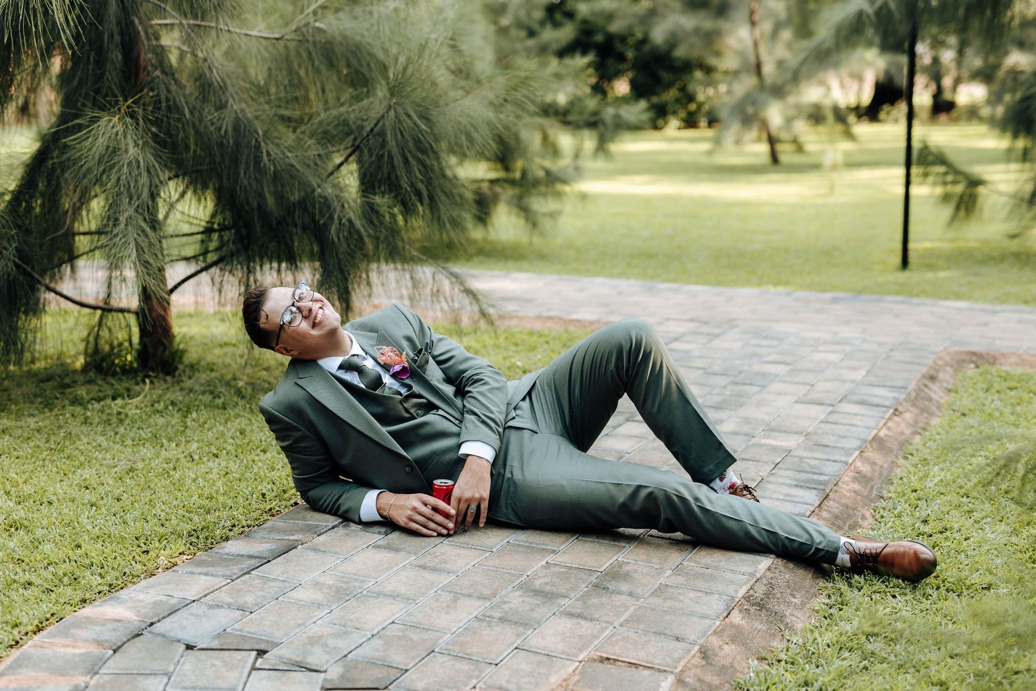 Person in green attire lounges casually on stone pavers under pine trees in a peaceful garden setting.