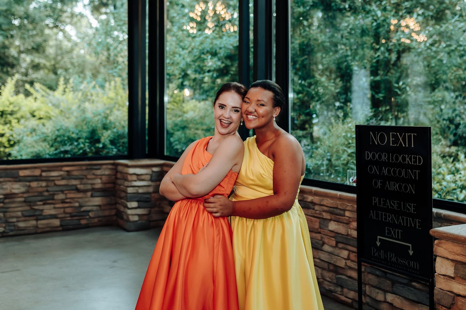 Two friends in vibrant orange and yellow dresses pose together on an outdoor patio.