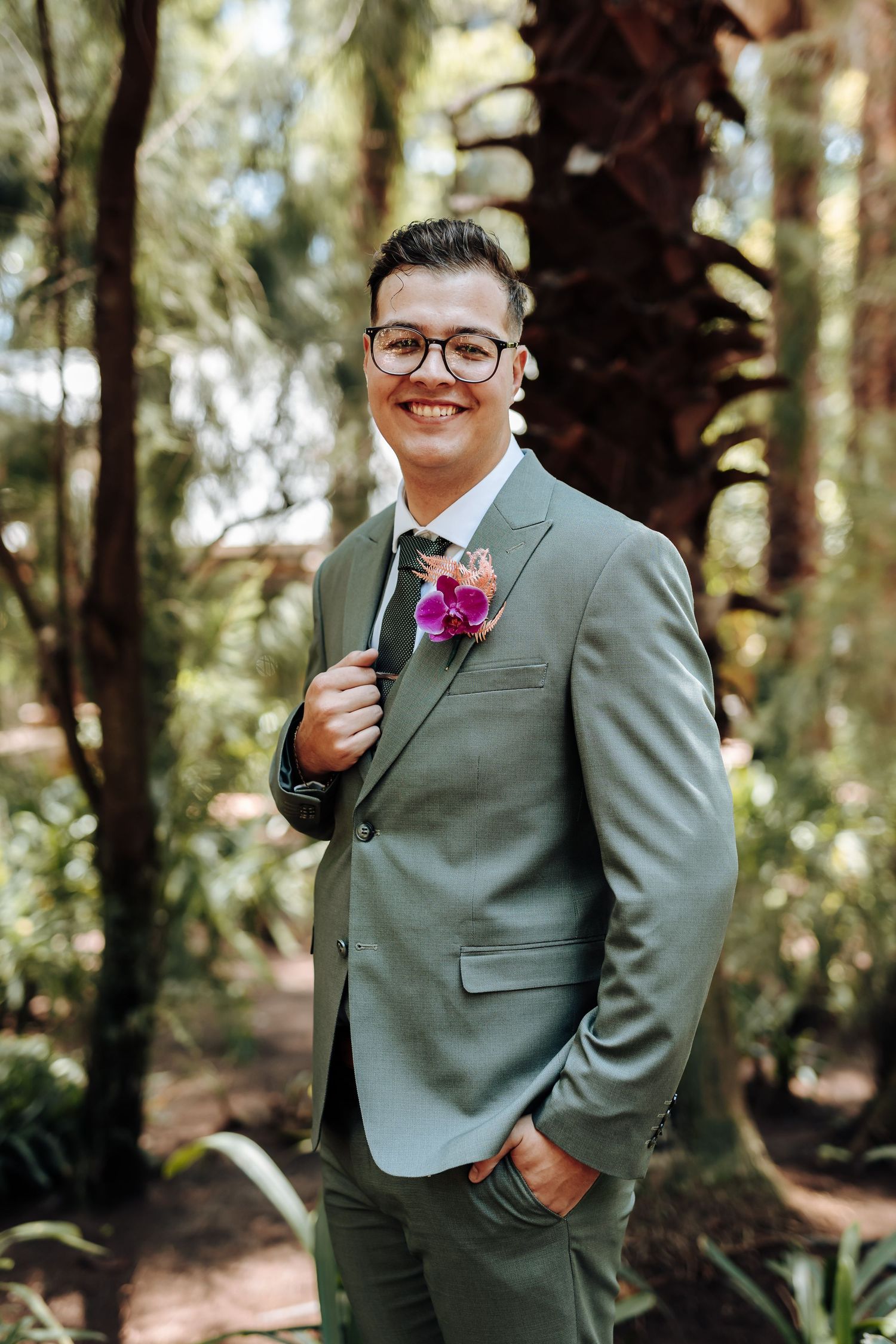 Wedding party member in a sage green suit poses confidently in a wooded outdoor setting with a purple boutonniere.