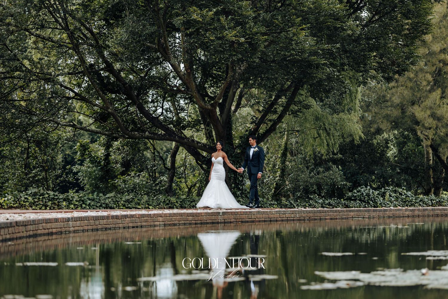 Wedding couple posing near reflective pond surrounded by lush greenery.