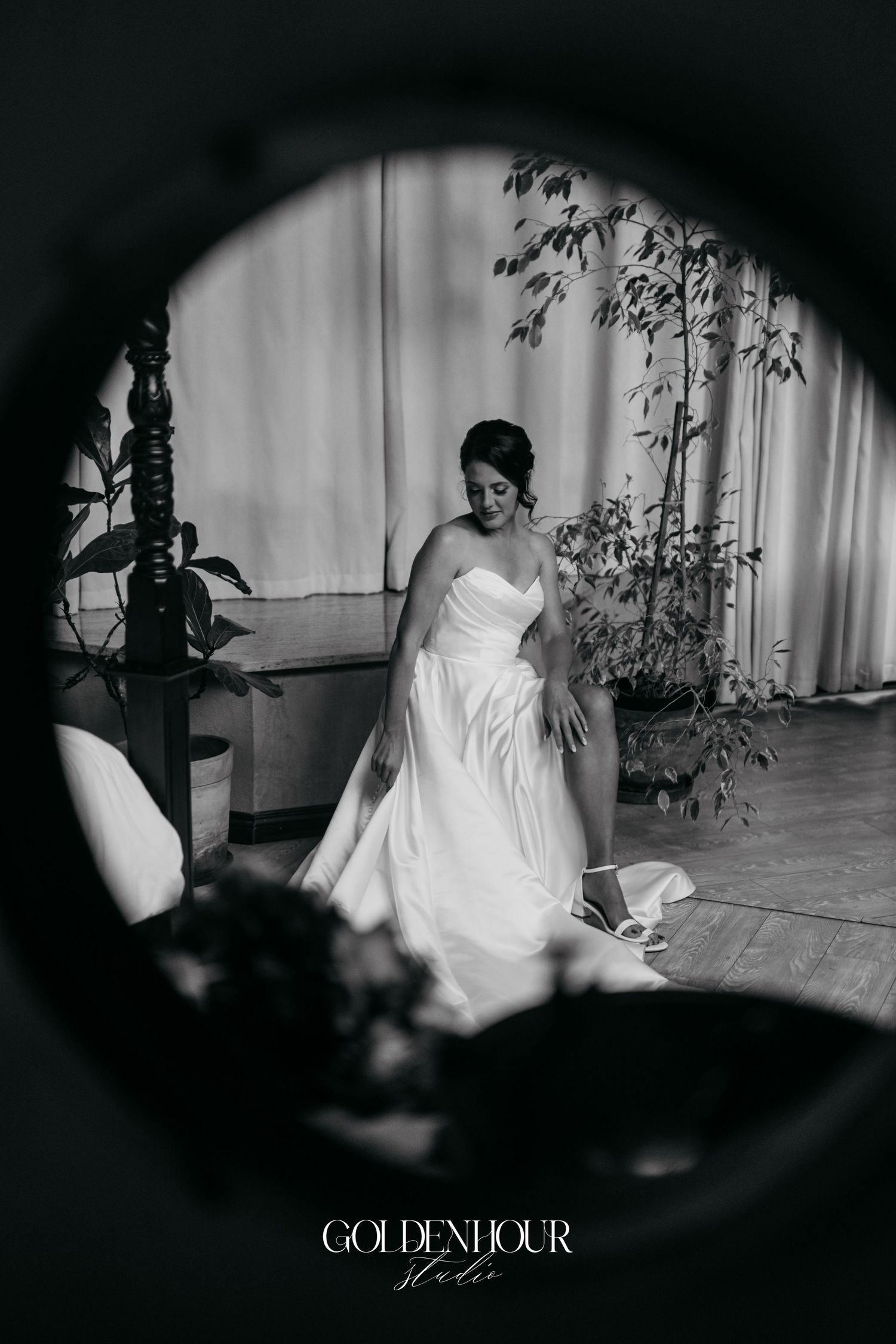 Black and white photo of a bride in a strapless gown reflected in a round mirror while adjusting her shoes.