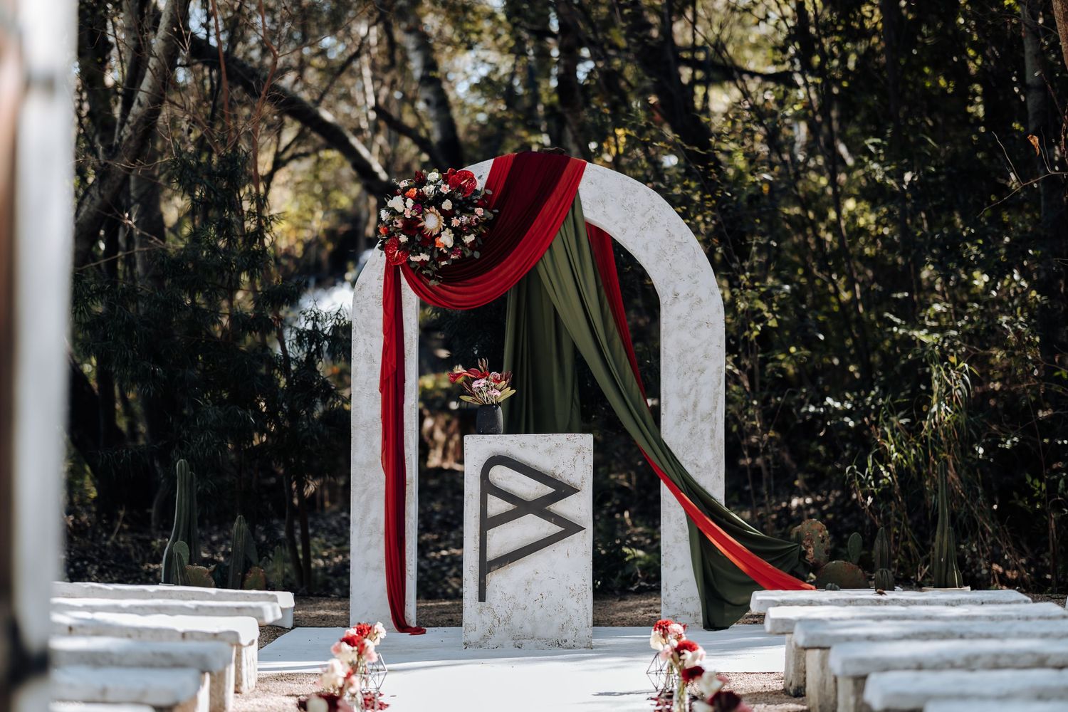 Elegant outdoor wedding arch adorned with red and green draped fabric and floral arrangements in a wooded setting.