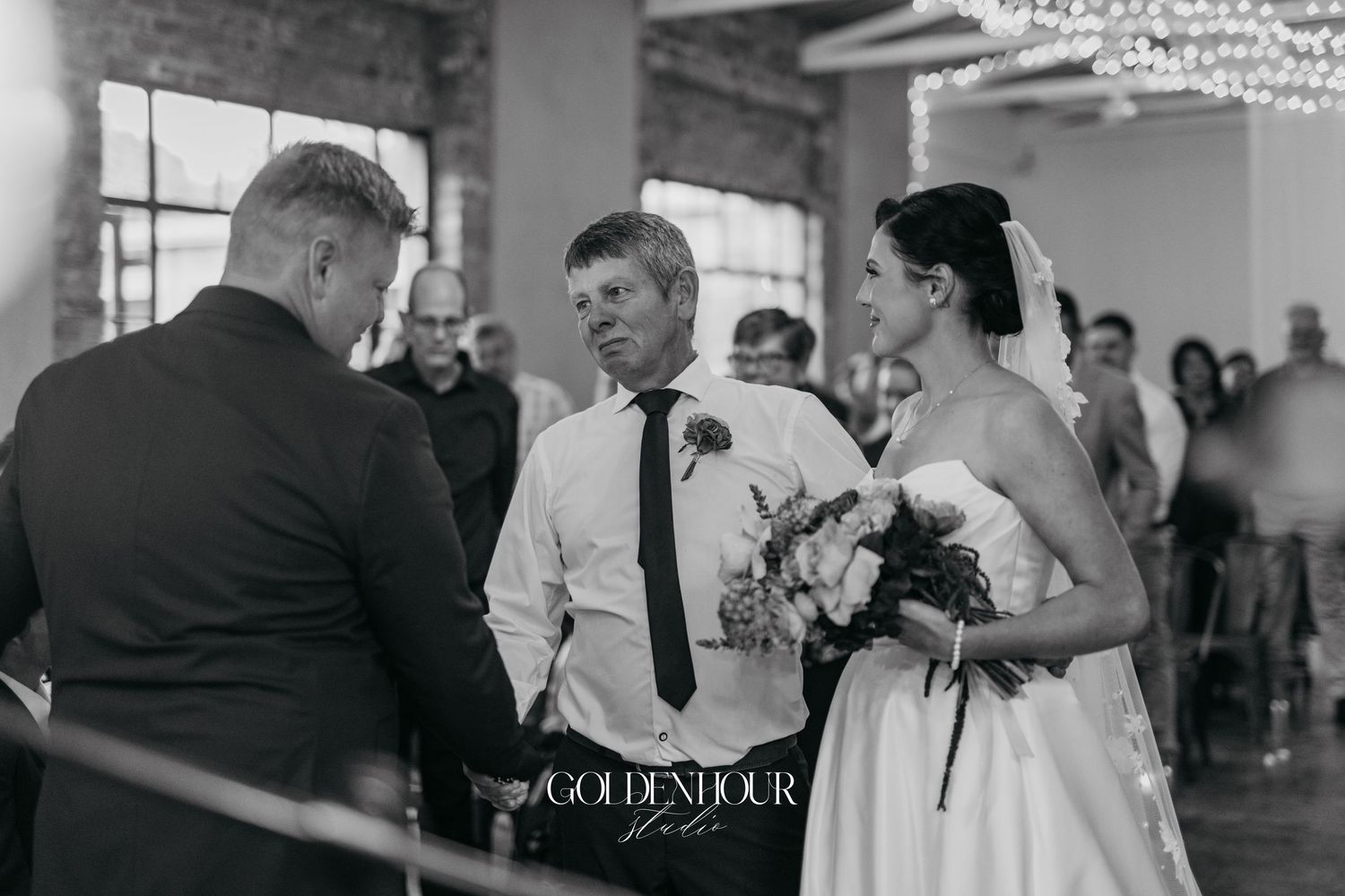 Black and white photo of a bride being walked down the aisle by her father during an indoor wedding ceremony.