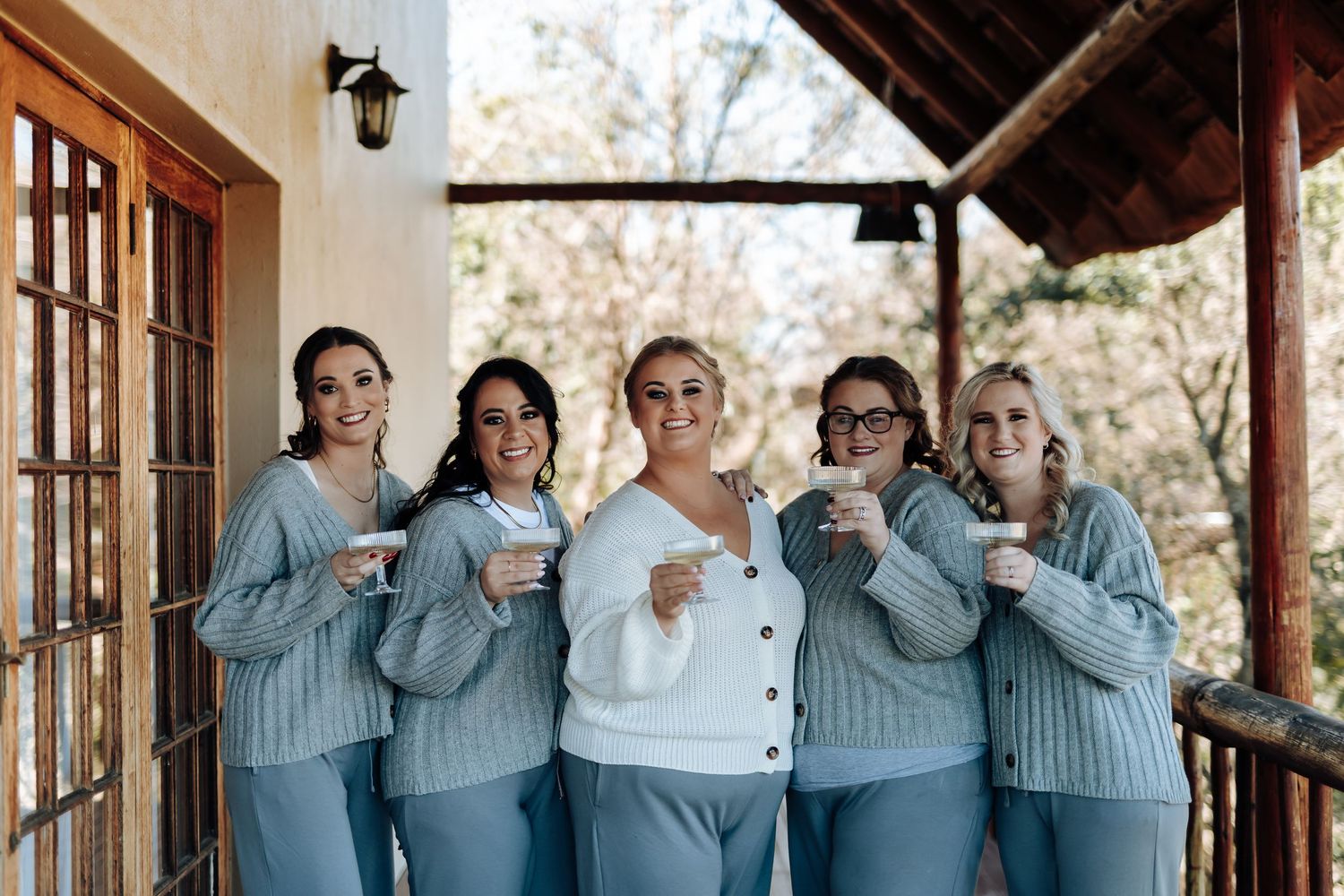 Five women in matching blue sweaters and pants smile holding mugs on a rustic porch, perfect for a bridal party.