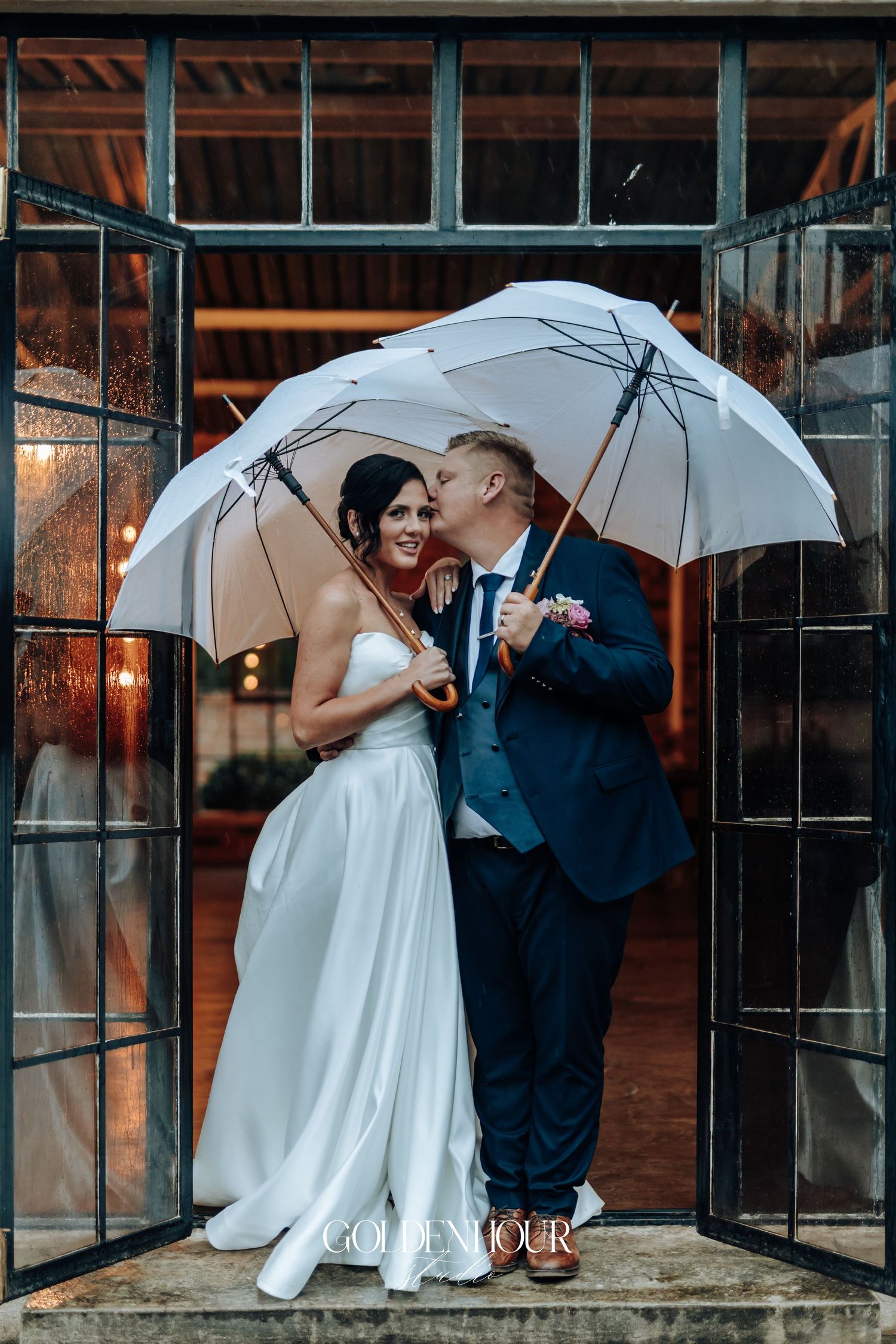 Bride and groom sharing a kiss under white umbrellas in the rain on their wedding day.