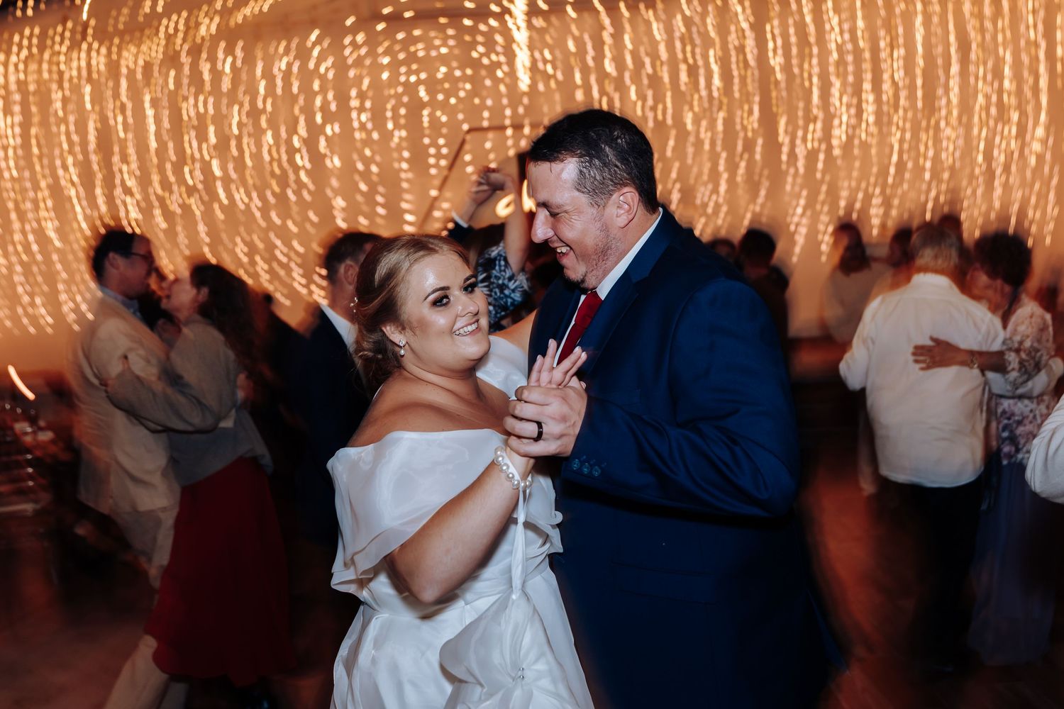 Bride and groom share first dance surrounded by warm fairy lights with wedding guests in background.