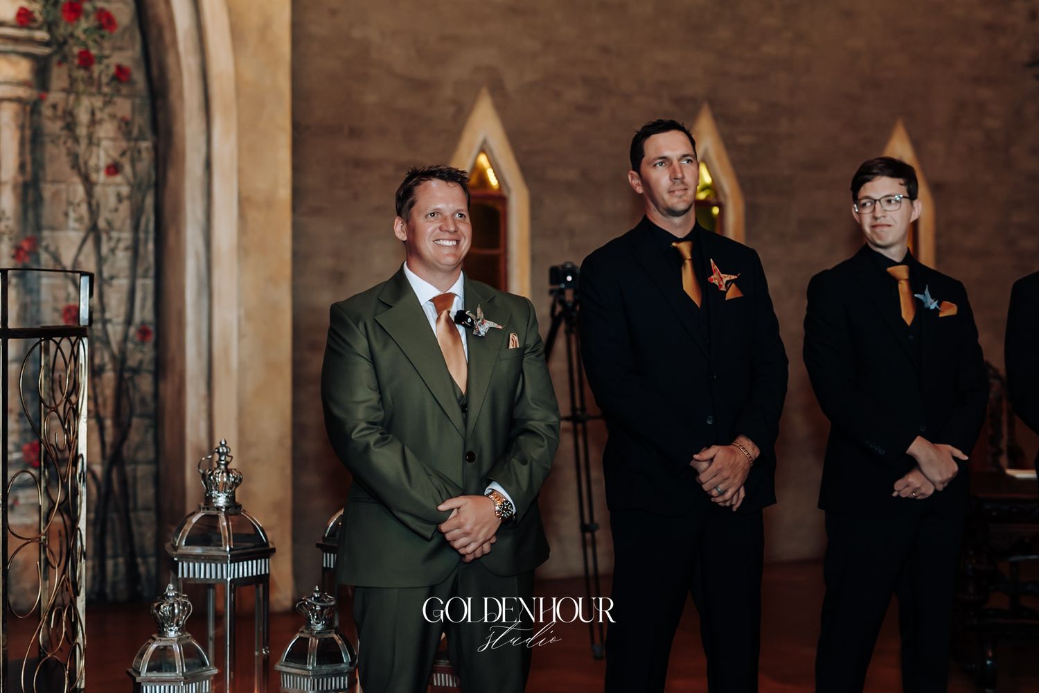 Groom in olive green suit with groomsmen in black standing at wedding ceremony altar.
