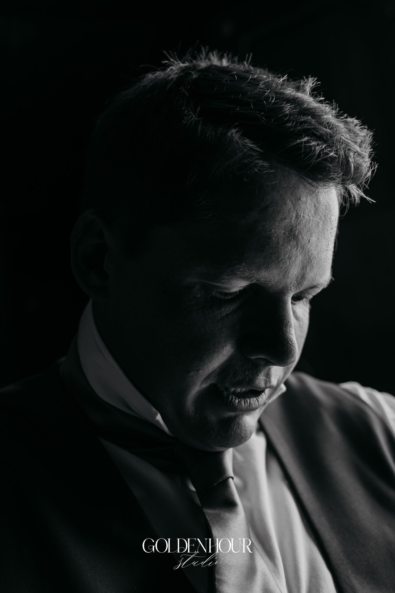 Dramatic black and white portrait of a groom in a suit looking down with moody lighting.
