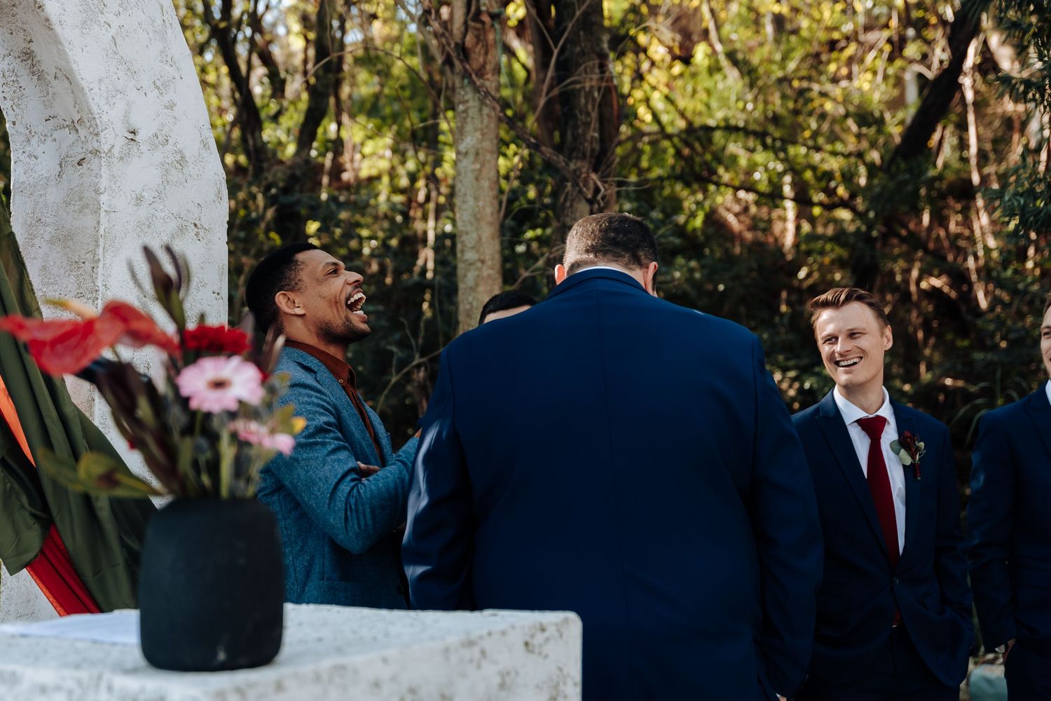 Three men in suits laughing together outdoors at a wedding ceremony with floral arrangements nearby.