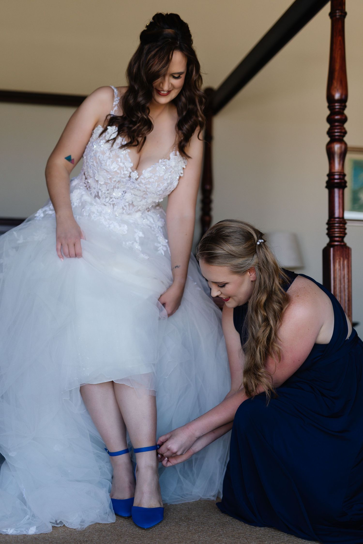 Helper in navy dress assists bride in white gown with putting on bright blue shoes during wedding preparations.