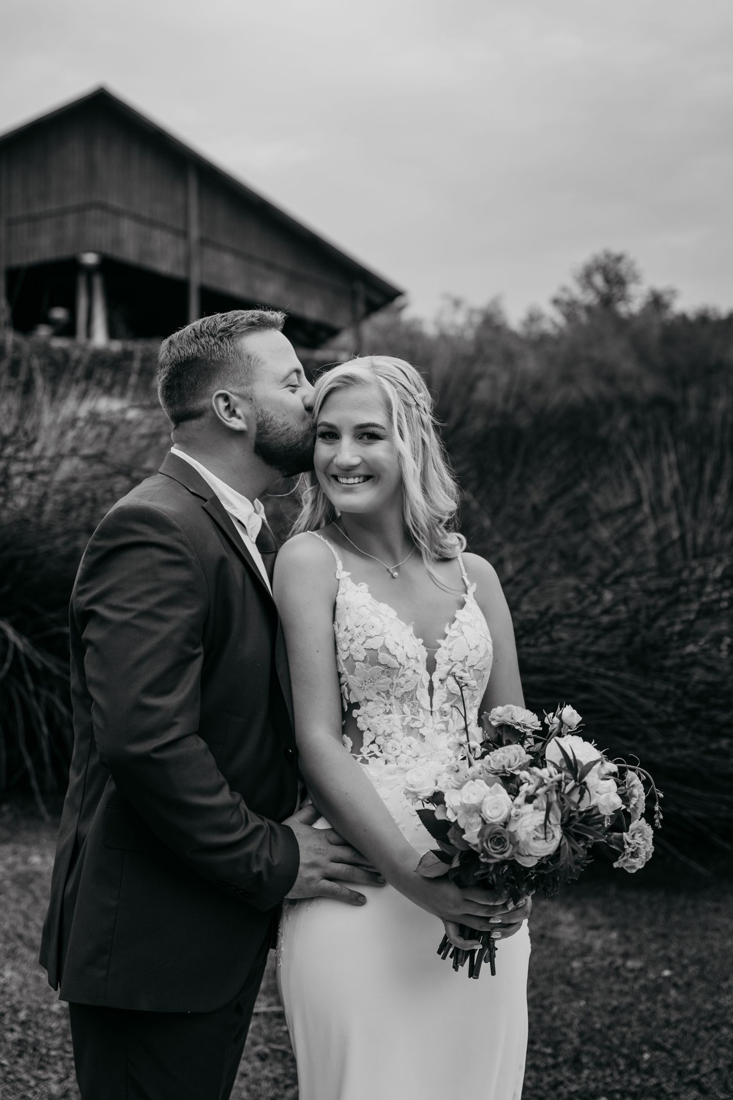 A romantic black and white wedding photo series showing tender moments between newlyweds in front of a rustic barn setting.