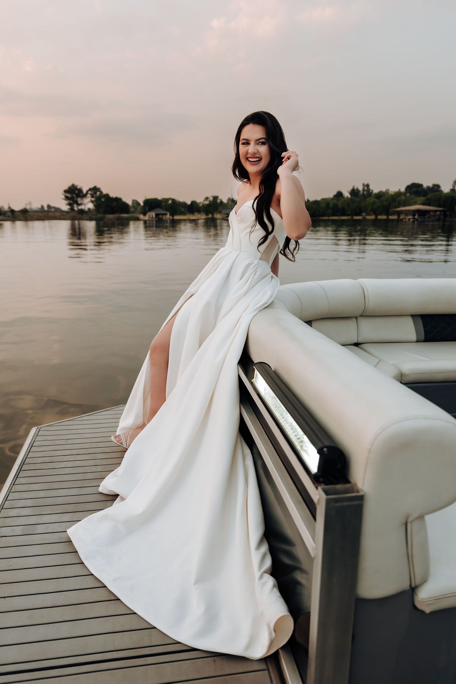 A bride in a classic white wedding gown poses elegantly on a boat deck at sunset over calm waters.