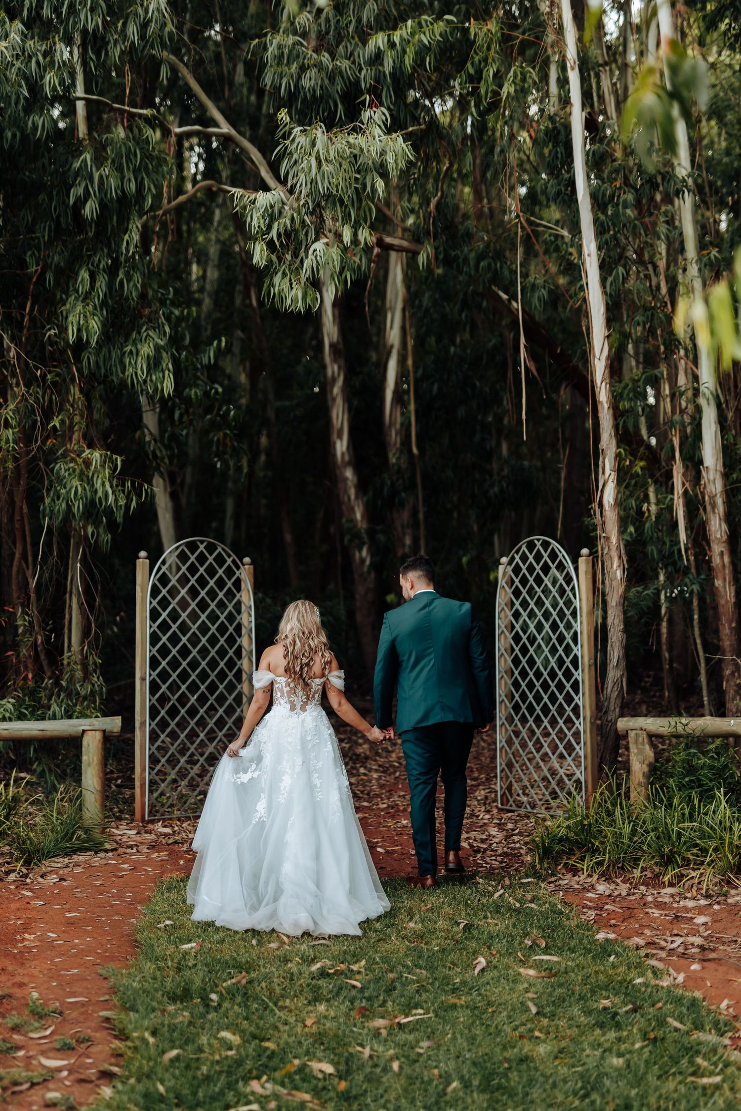 A couple in wedding attire walks away into a eucalyptus forest path with decorative lattice panels.